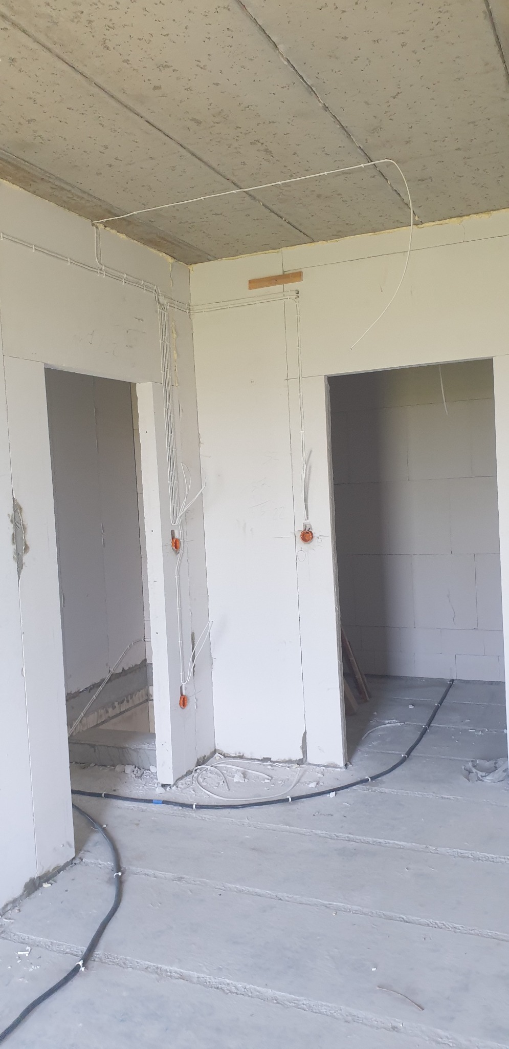 Surowy stan wnętrza z widocznymi przewodami elektrycznymi poprowadzonymi po ścianach i suficie, pomarańczowe puszki pod gniazdka, czarny kabel na podłodze.