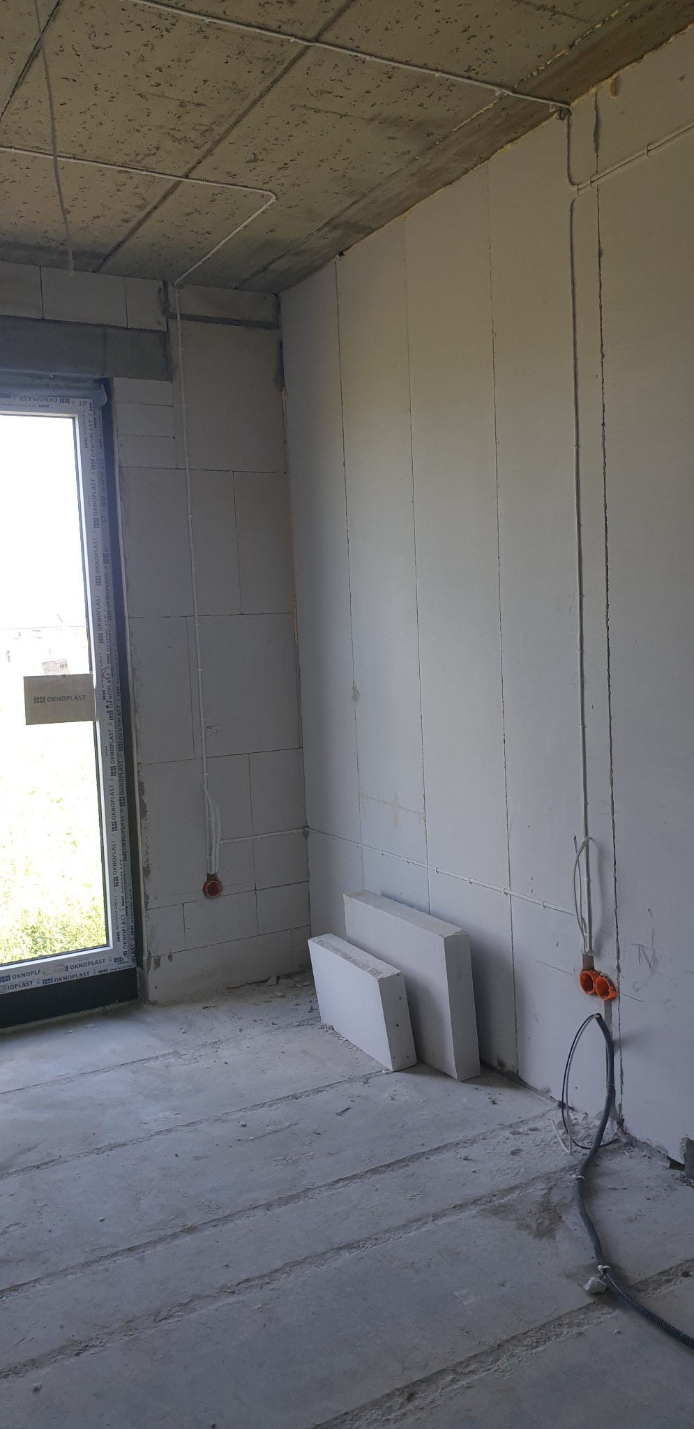 Surowy stan wnętrza z widoczną instalacją elektryczną w rurkach na ścianie, puszkami elektrycznymi w murze i przewodami na podłodze, widok na okno z szybą z logo producenta.