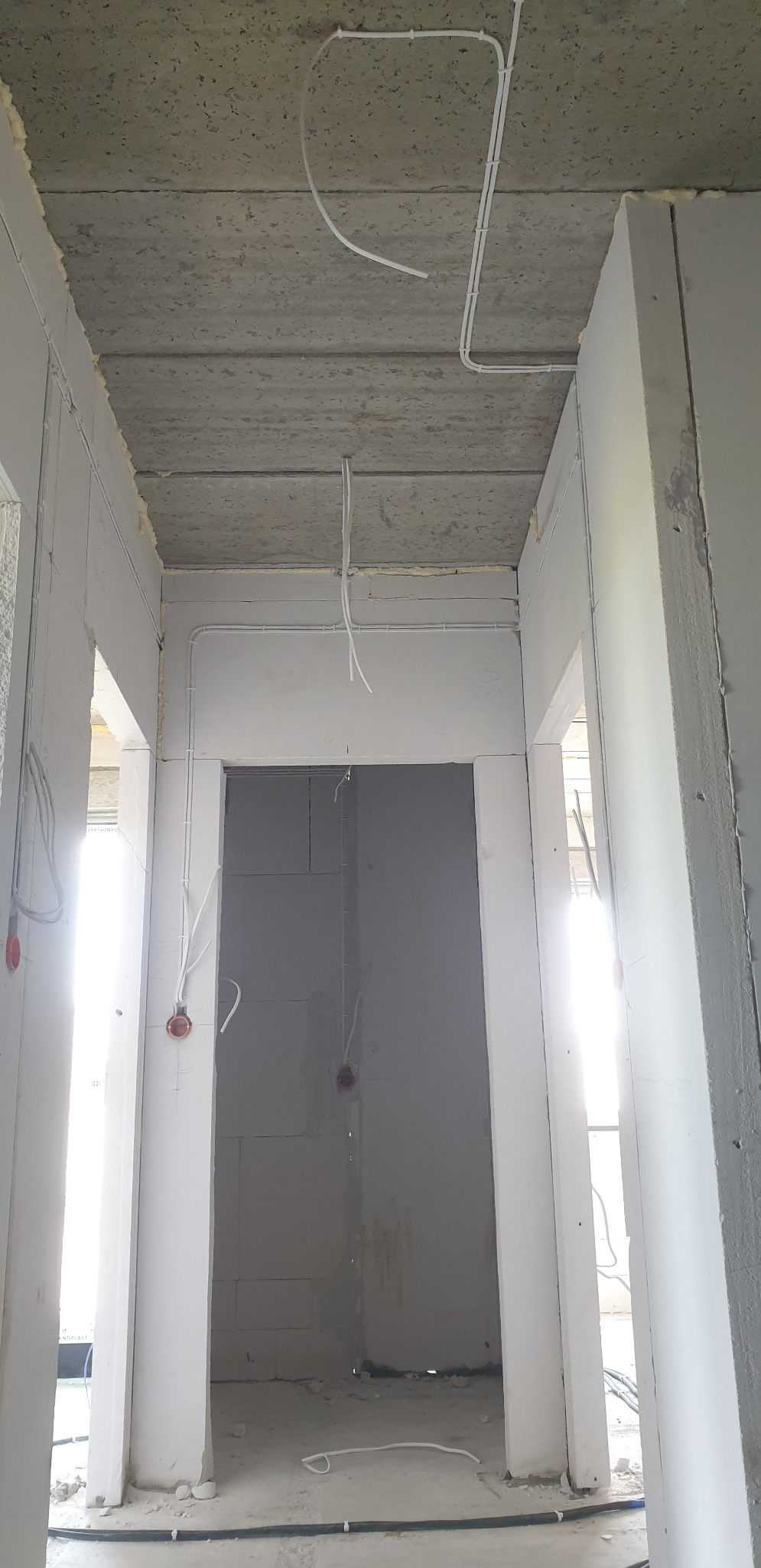 Wnętrze w trakcie budowy z widocznymi przewodami elektrycznymi poprowadzonymi po suficie i ścianach, z puszkami instalacyjnymi w kolorze pomarańczowym, surowy betonowy strop i białe ściany z pustaków.