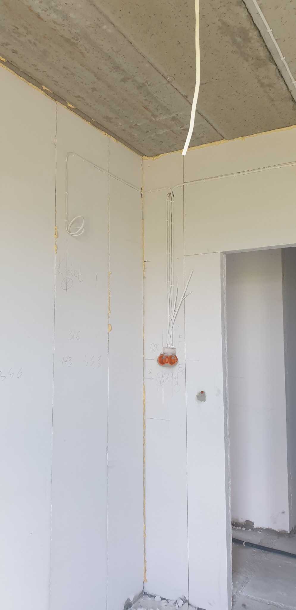 Surowy stan wnętrza z widocznymi przewodami elektrycznymi wychodzącymi ze ściany, pomarańczową puszką instalacyjną i szkicem technicznym na ścianie, ukazujący etap prac wykończeniowych.