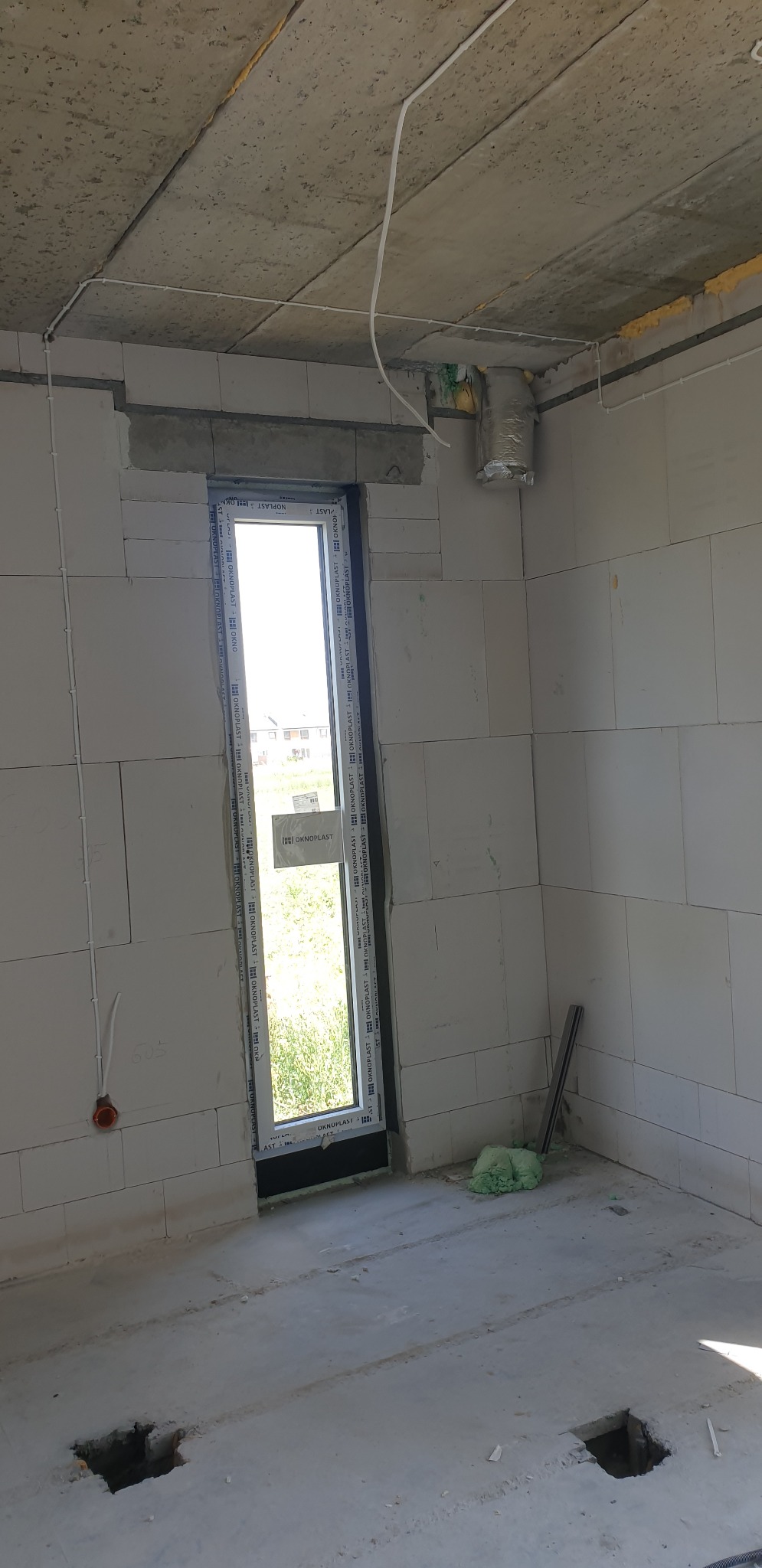 Surowy stan wnętrza budynku w trakcie budowy; widoczne przewody pod sufitem, otwór w ścianie z oknem, oraz otwory w podłodze