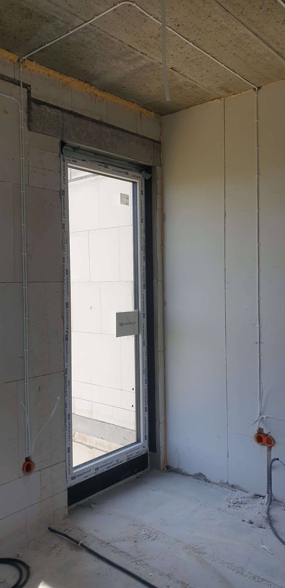 Surowy stan wnętrza z widocznymi przewodami elektrycznymi poprowadzonymi po ścianach i suficie, pomarańczowe puszki instalacyjne w ścianie, okno z zamontowaną roletą zewnętrzną, widoczne elementy...