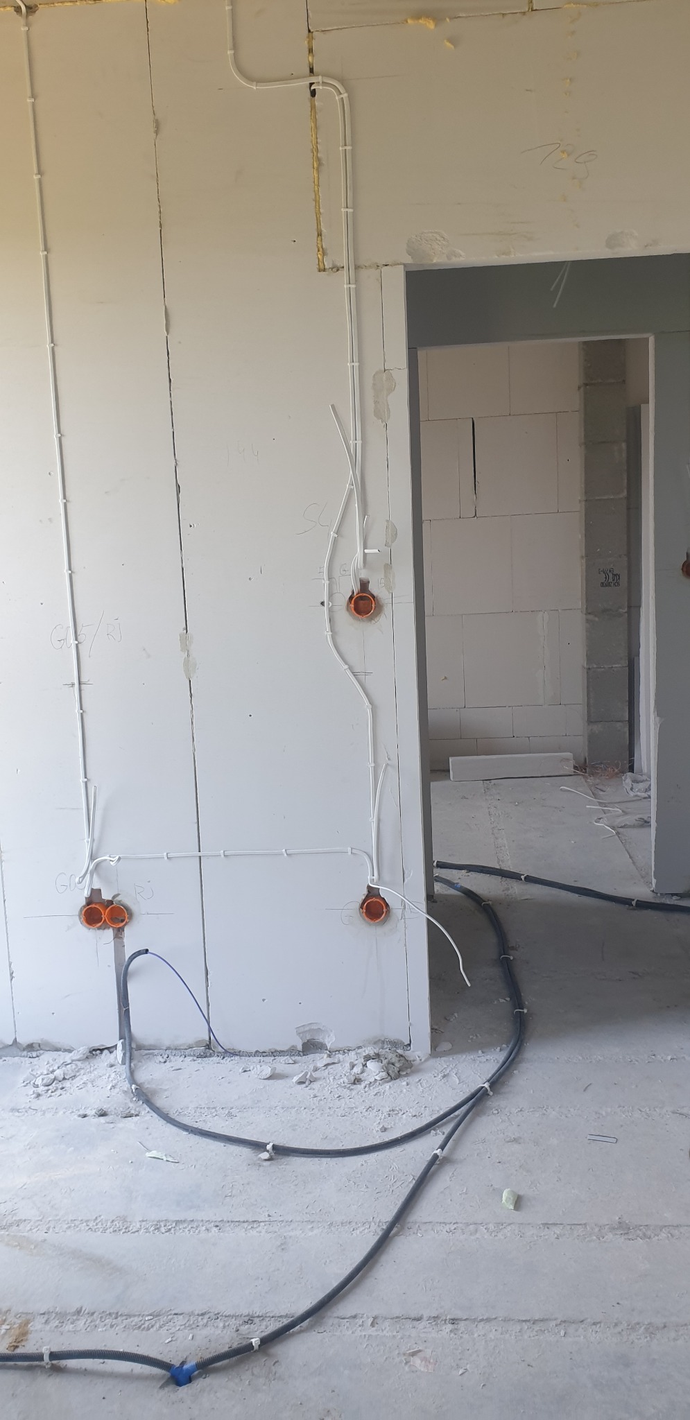 Wnętrze w trakcie prac wykończeniowych; widoczne białe ściany z zamontowanymi puszkami elektrycznymi i poprowadzonymi przewodami w białych listwach oraz czarne kable na podłodze w surowym stanie.