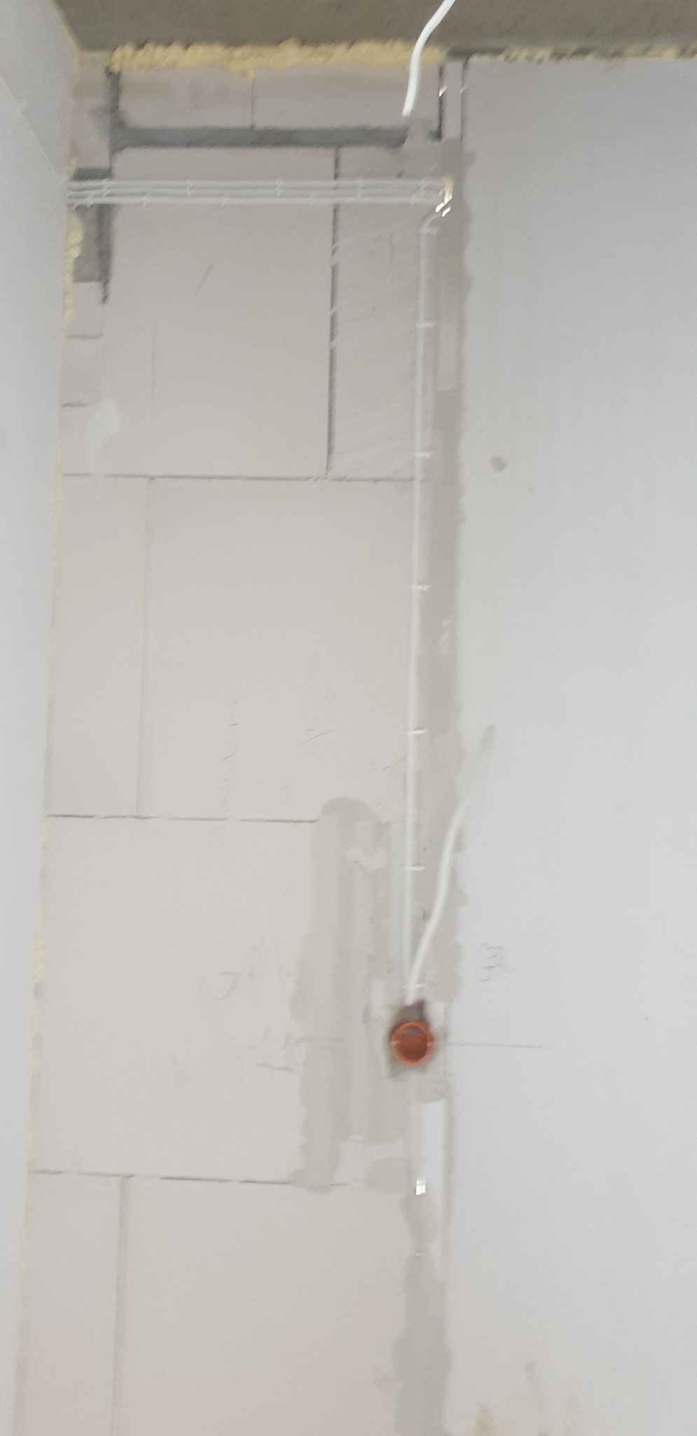 Surowy stan: biała ściana z pustaków z widocznymi przewodami w peszlu i zamontowaną pomarańczową puszką pod gniazdko elektryczne.