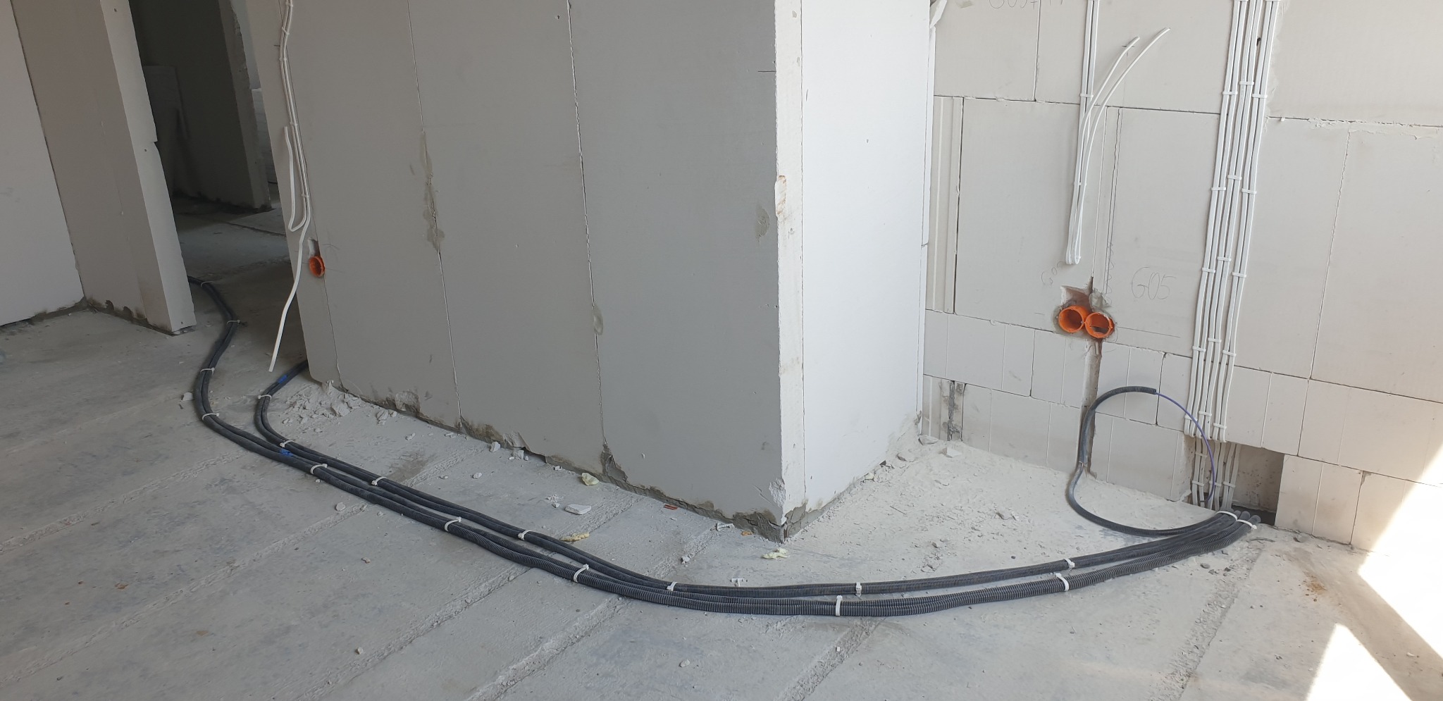 Cztery czarne rury karbowane ułożone równolegle na surowym betonowym podłożu, prowadzone wzdłuż ściany z białych bloczków, z widocznymi puszkami instalacyjnymi i przewodami w ścianie.