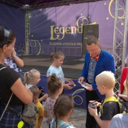 Iluzjonista w niebieskiej marynarce i muszce w kropki prezentuje karty z trikami grupie dzieci na tle sceny z napisem 'Legend' i 'Śląskie Wesołe Mia'