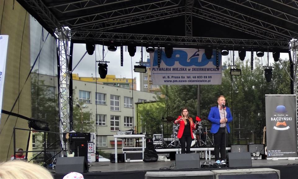 Występ duetu na scenie plenerowej: mężczyzna w niebieskiej marynarce z muszką i kobieta w czerwonej marynarce, za nimi perkusja i baner Pływalni Miejskiej w Skierniewicach.