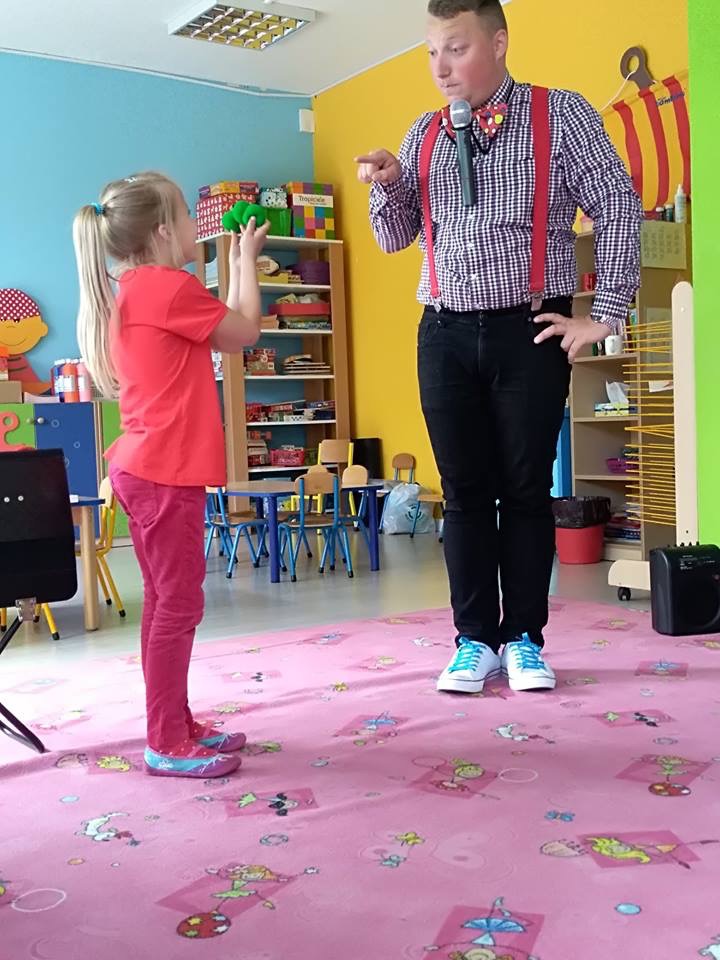 Mężczyzna w szelkach i muszce, z mikrofonem w dłoni, pokazuje palcem na dziewczynkę trzymającą zielony przedmiot w sali zabaw.