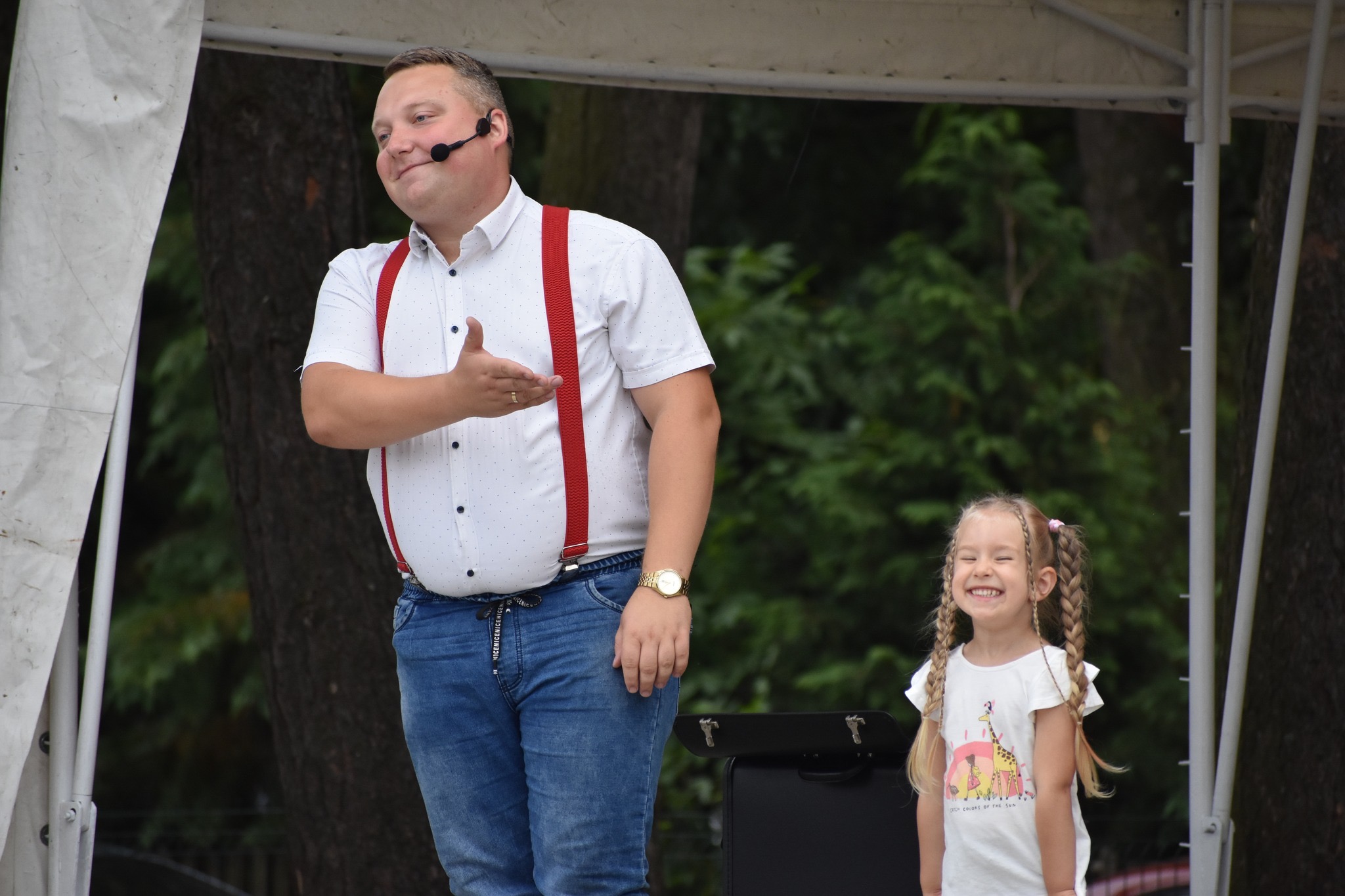 Uśmiechnięty iluzjonista w czerwonych szelkach i mikrofonem na głowie prezentuje się obok roześmianej dziewczynki na scenie podczas występu plenerowego.