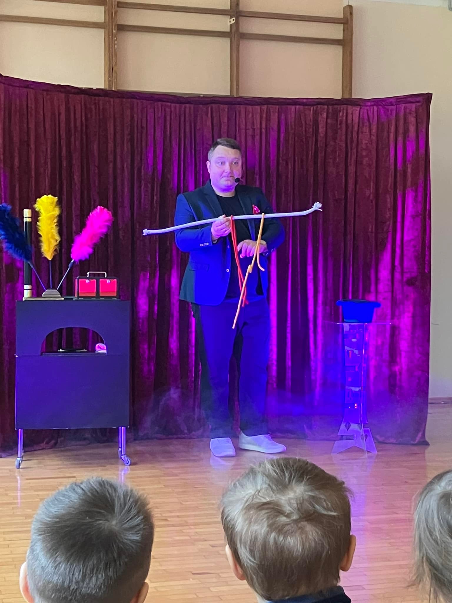 Iluzjonista w czarnym garniturze prezentuje trik z linami na scenie z bordową zasłoną w tle, oświetlony fioletowymi światłami, widownia z przodu.