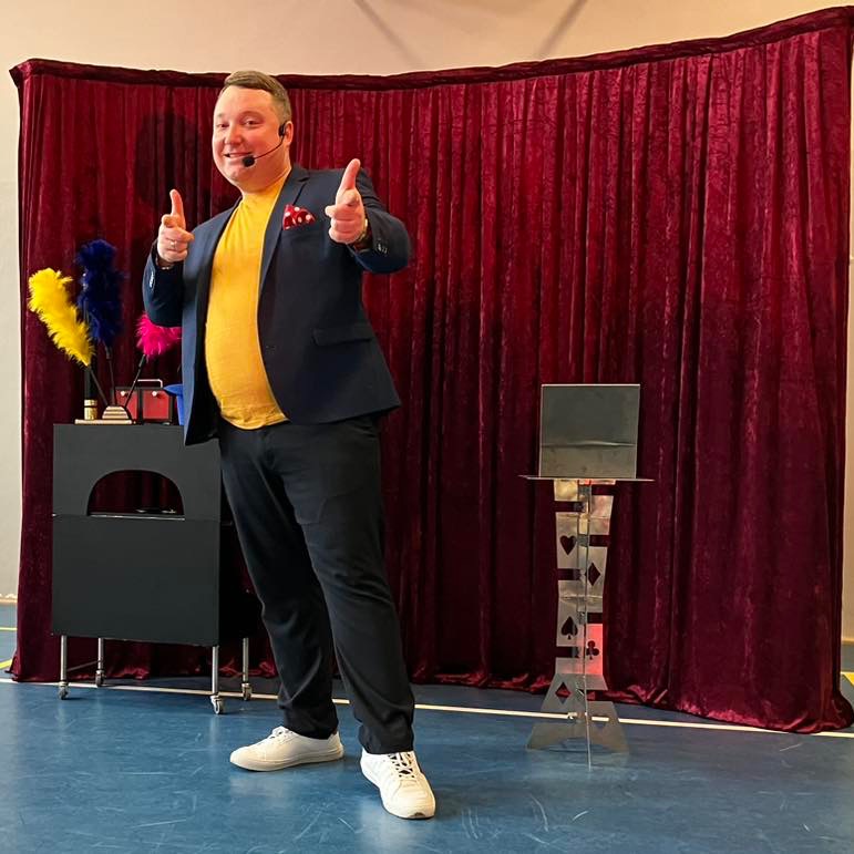 Uśmiechnięty iluzjonista w niebieskiej marynarce i żółtej koszulce, wskazujący palcami w stronę widza, na scenie z czerwonymi zasłonami i metalowym stolikiem z kartami.