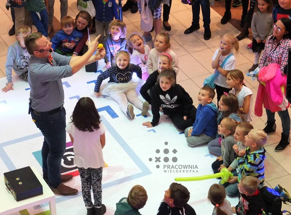 Iluzjonista w koszuli w kratę i muszce prezentuje sztuczkę z żółtą piłeczką grupie dzieci siedzących na podłodze z nadrukowanym logo Pracownia Wielokropka.