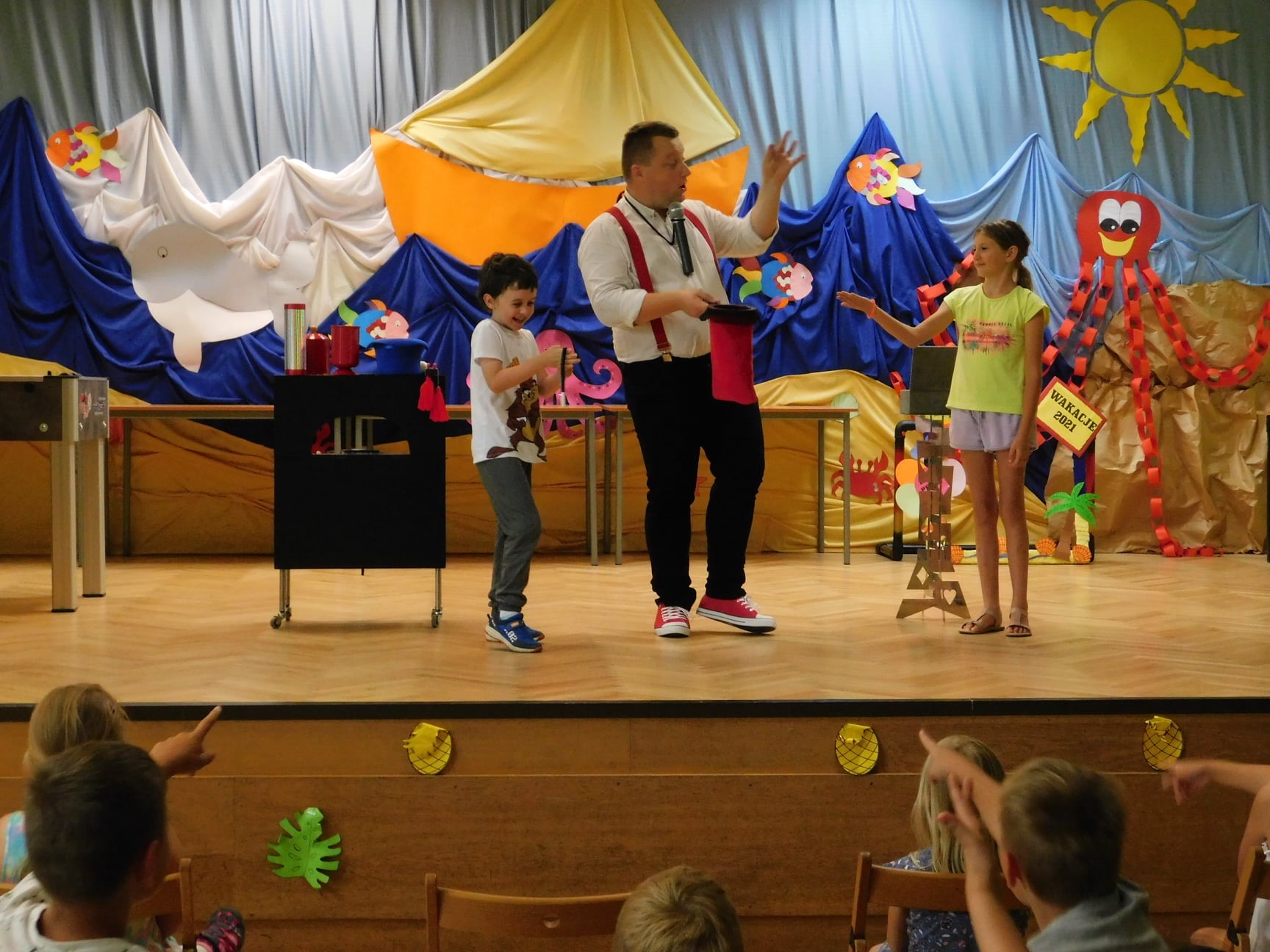 Magik w czerwonych szelkach prezentuje trik z czerwoną chustą i czarnym cylindrem na scenie z dziećmi podczas występu o tematyce wakacyjnej, z dekoracjami morskimi w tle.