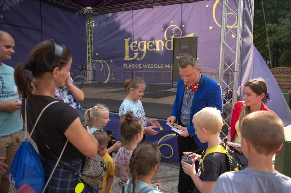 Iluzjonista w niebieskiej marynarce i muszce w kropki prezentuje karty z trikami grupie dzieci na tle sceny z napisem 'Legend' i 'Śląskie Wesołe Mia'
