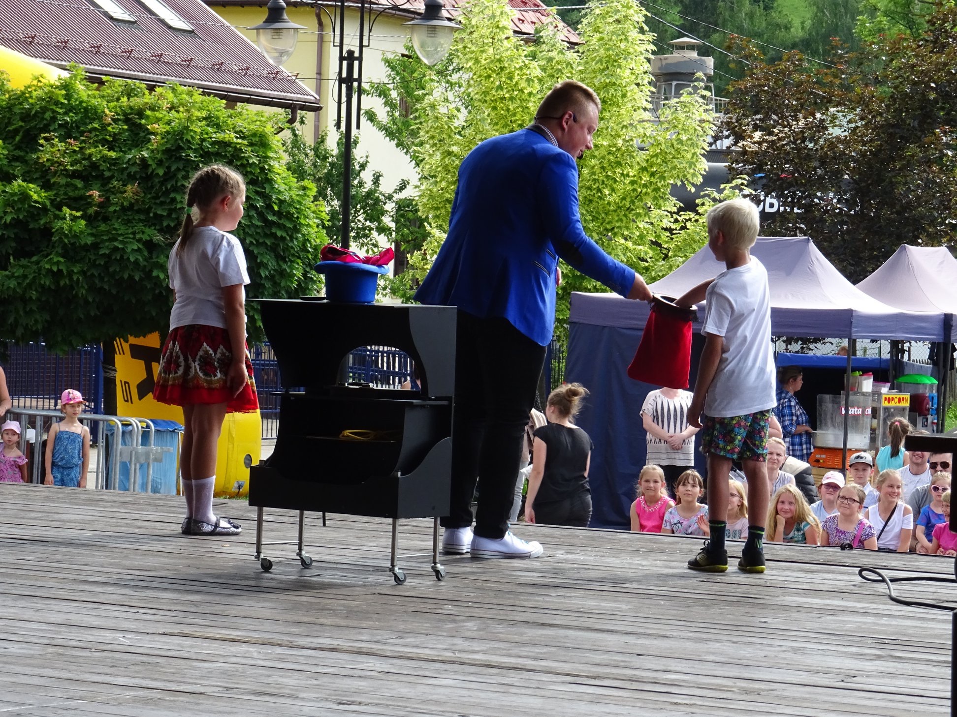 Iluzjonista w niebieskiej marynarce prezentuje sztuczkę z czerwoną torbą na drewnianej scenie, obserwuje go blond chłopiec i dziewczynka w czerwonej spódnicy oraz zgromadzona publiczność.