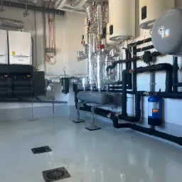 Nowoczesna kotłownia z białymi bojlerami, srebrnymi rurami izolowanymi i czarnymi rurami instalacji. Widoczne filtry i zawory. Podłoga z odpływami.