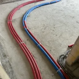 Instalacja ogrzewania podłogowego w trakcie montażu: czerwone i niebieskie rury ułożone na surowym betonie, zabezpieczone srebrnymi klamrami.
