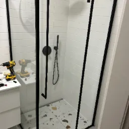 Nowoczesna kabina prysznicowa z czarnymi profilami, białe płytki i terazzo. W tle wiertarki sugerują montaż w trakcie realizacji.