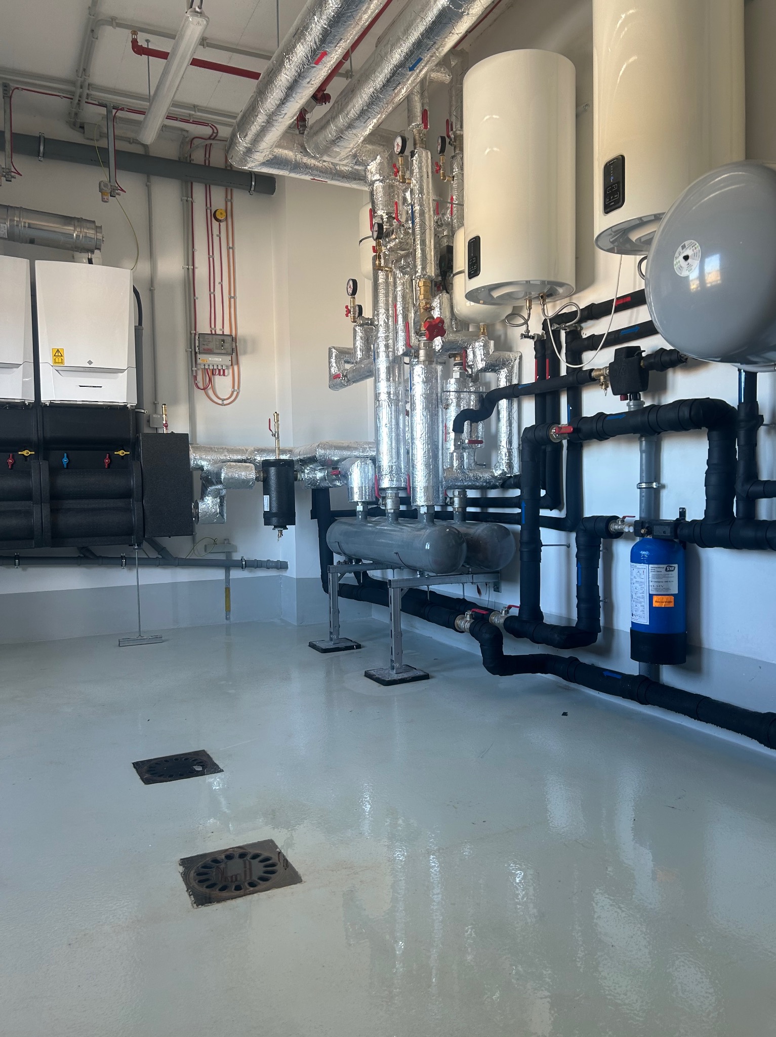 Nowoczesna kotłownia z białymi bojlerami, srebrnymi rurami izolowanymi i czarnymi rurami instalacji. Widoczne filtry i zawory. Podłoga z odpływami.