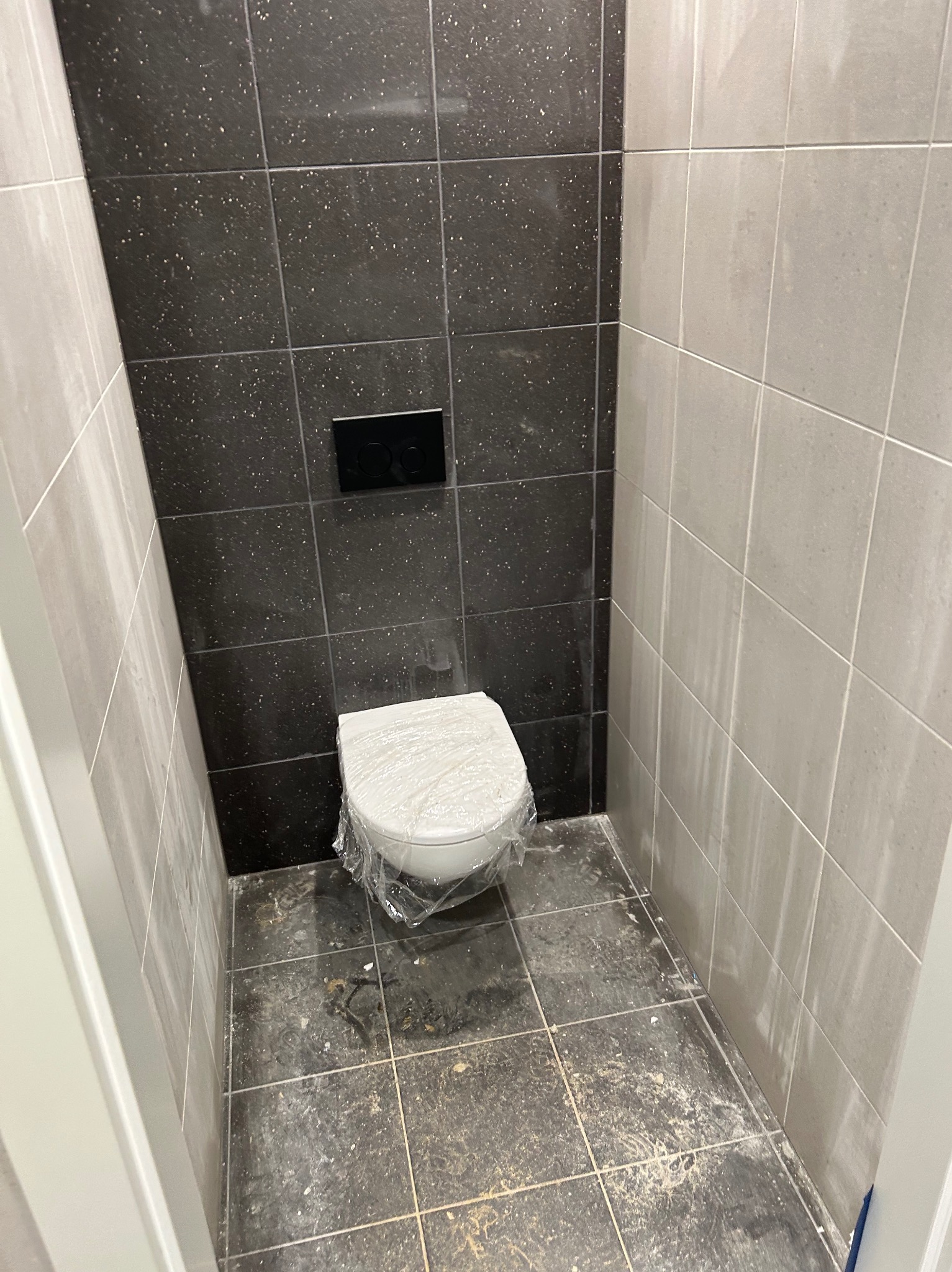 Wnętrze toalety po remoncie. Widoczne czarne i szare płytki na ścianach, podwieszana muszla WC zabezpieczona folią i czarny przycisk spłuczki. Ujęcie z poziomu podłogi.