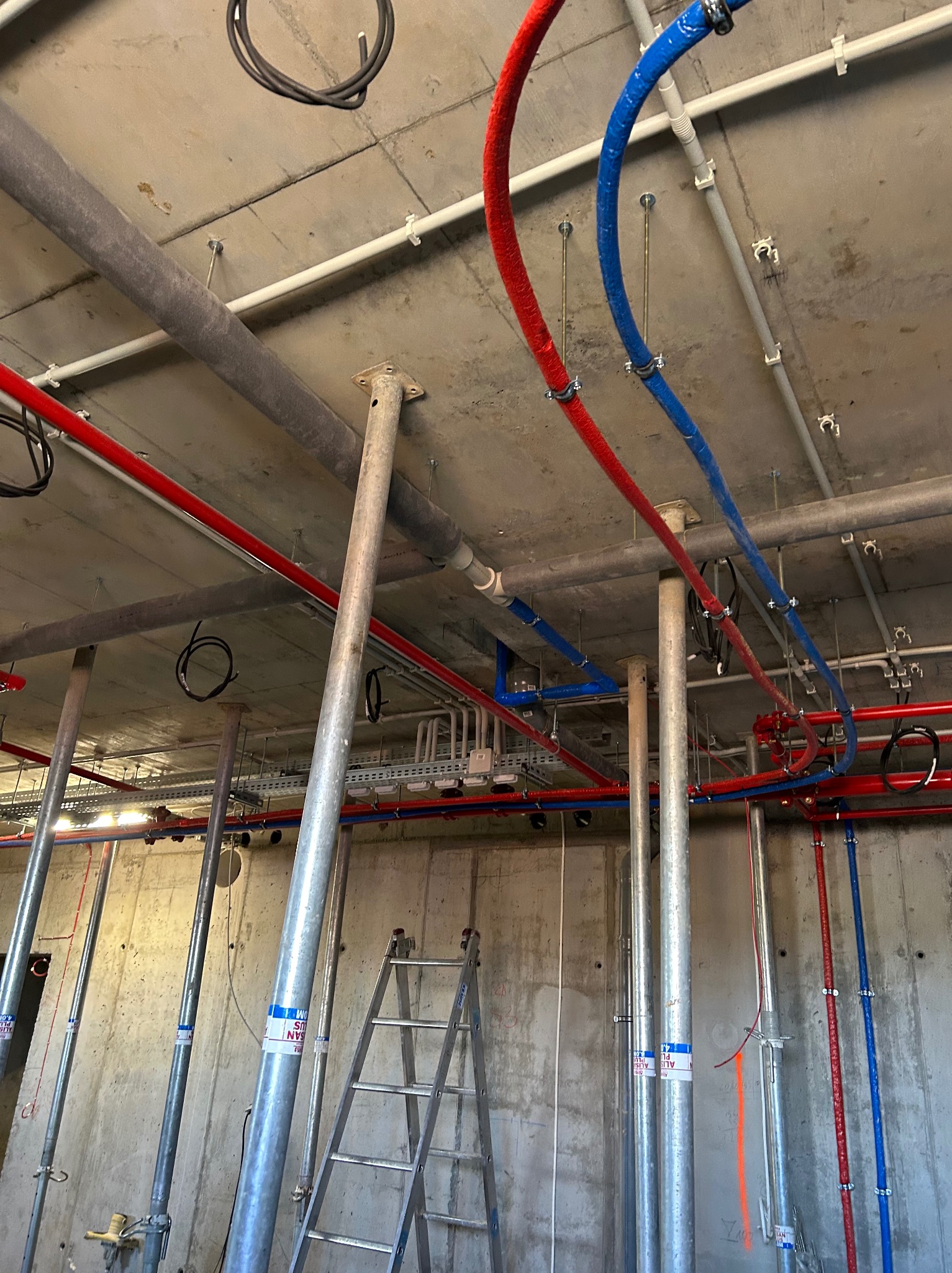 Instalacja rur grzewczych (czerwone i niebieskie) pod sufitem w surowym stanie, widoczne wsporniki i drabina. Instalacje w budynku w trakcie budowy.