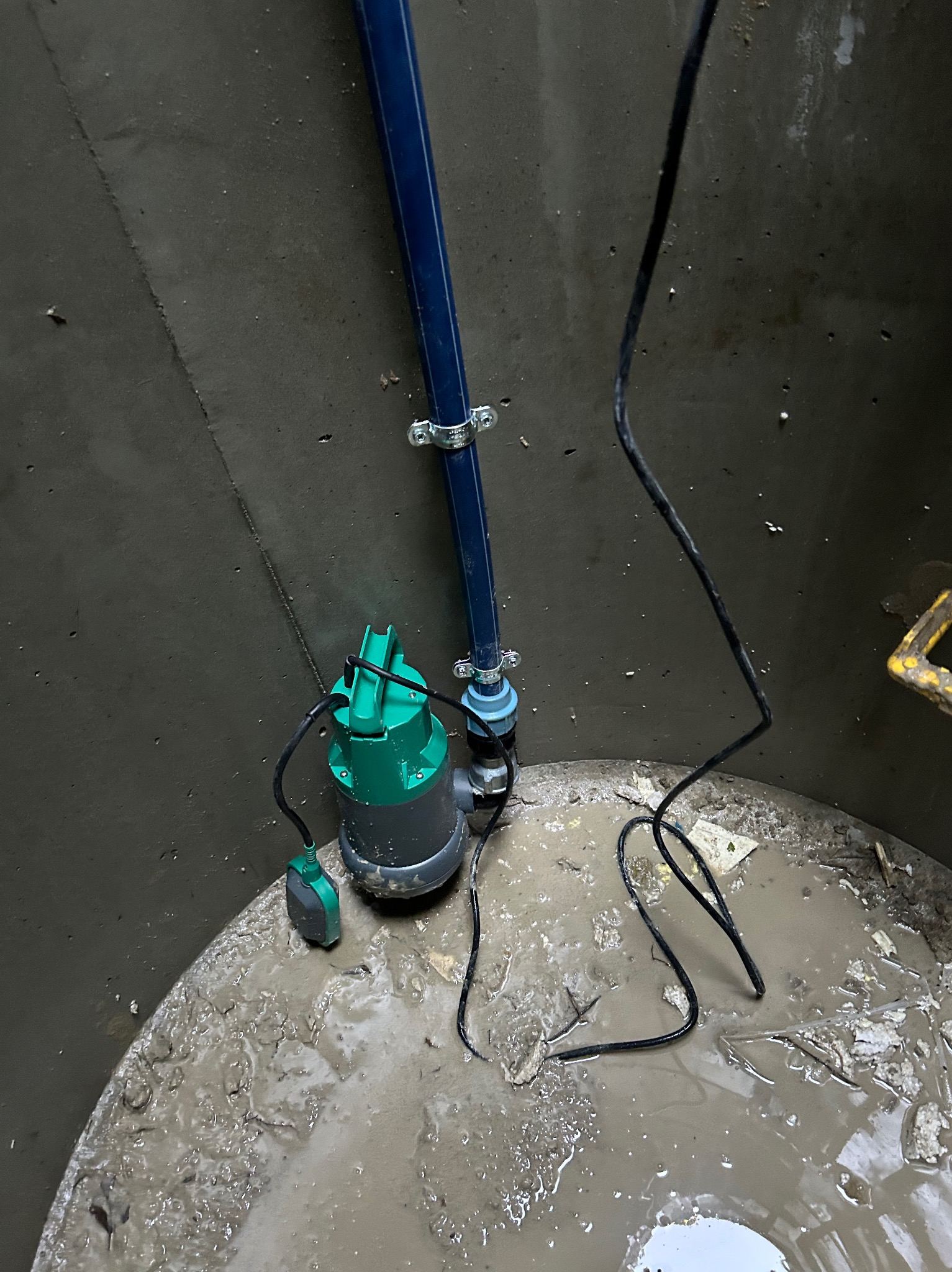 Zanurzona pompa z zieloną obudową i niebieską rurą w szambie betonowym wypełnionym brudną wodą. Widoczne kable i elementy instalacji.