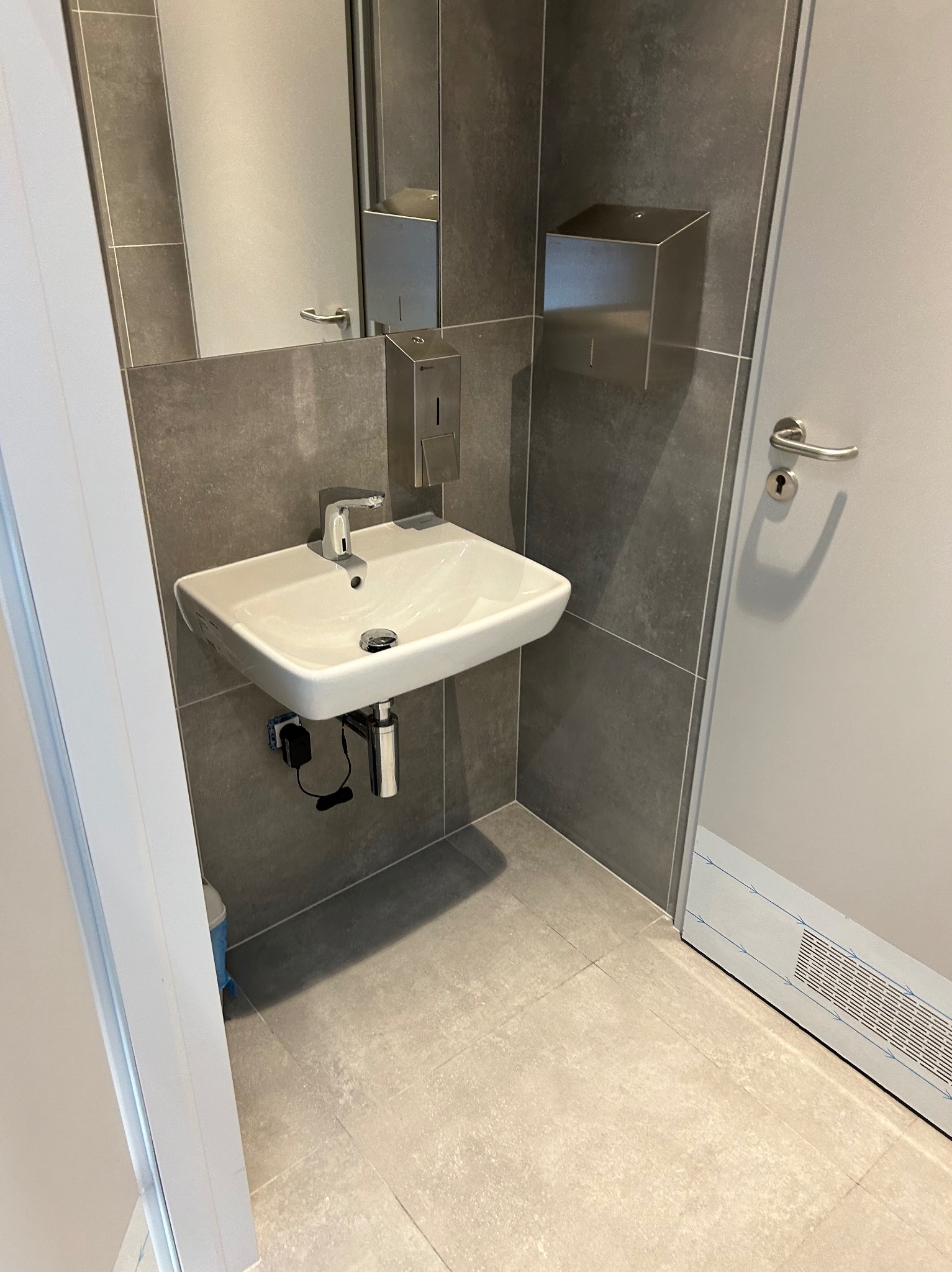 Nowoczesna łazienka z białą umywalką, chromowaną armaturą i szarą glazurą. Widoczny dozownik mydła i fragment drzwi z wentylacją.