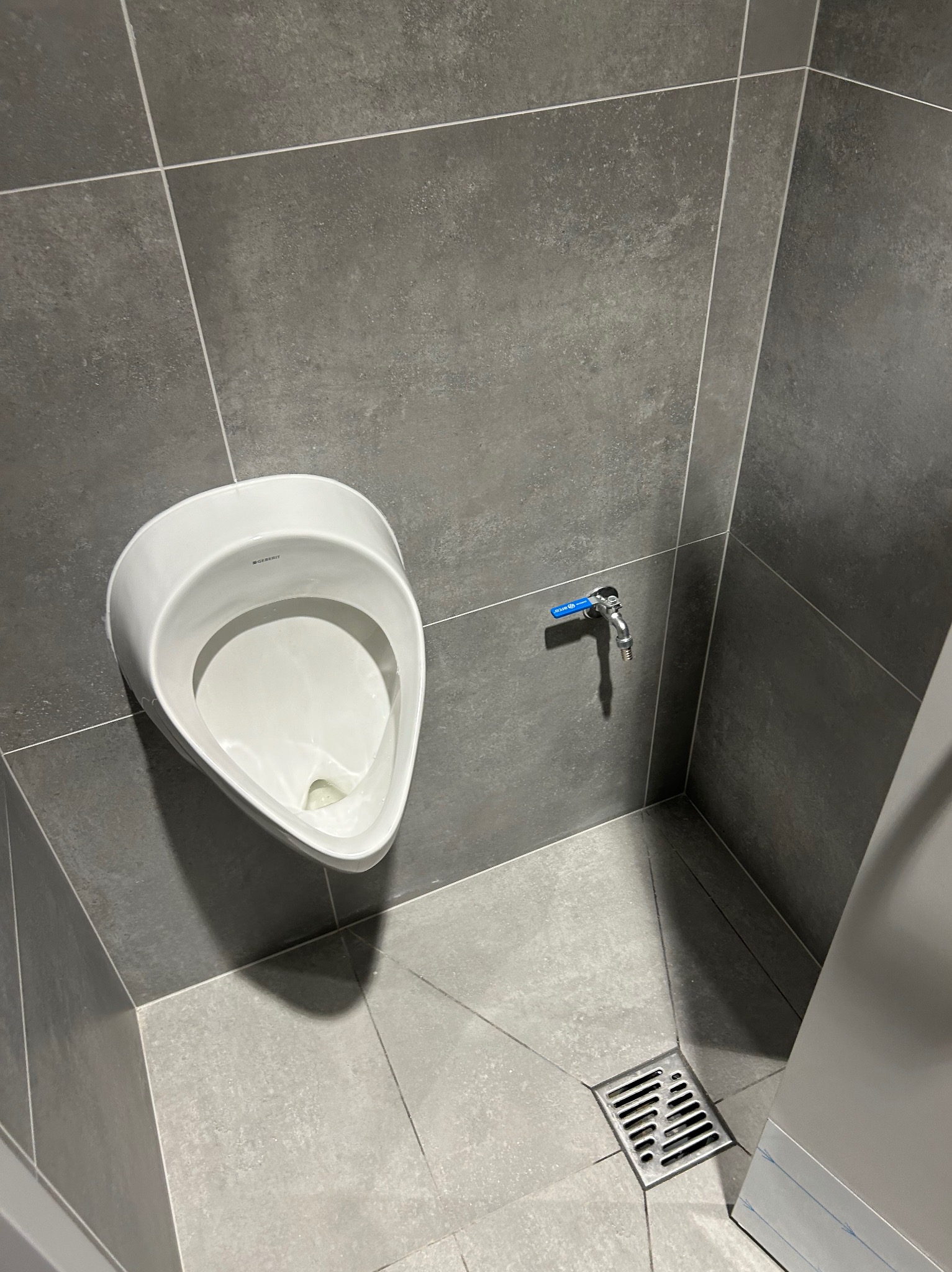 Biały pisuar marki Geberit na szarej, wykafelkowanej ścianie i podłodze. Widoczny kran z niebieskim kurkiem i odpływ podłogowy.
