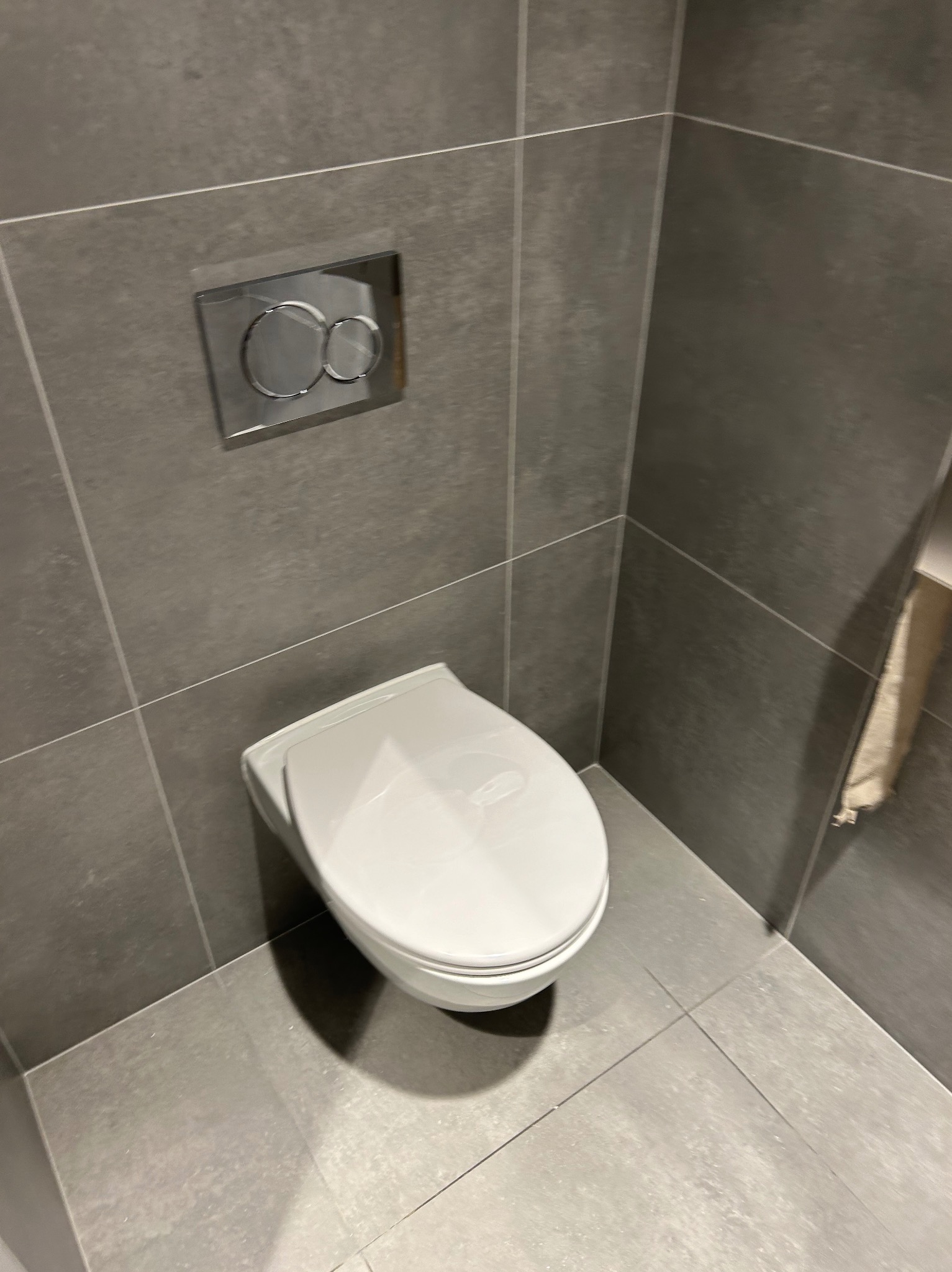 Nowoczesna toaleta podwieszana z chromowanym przyciskiem spłukującym na tle szarych płytek. Minimalistyczny design łazienki z ręcznikiem w tle.