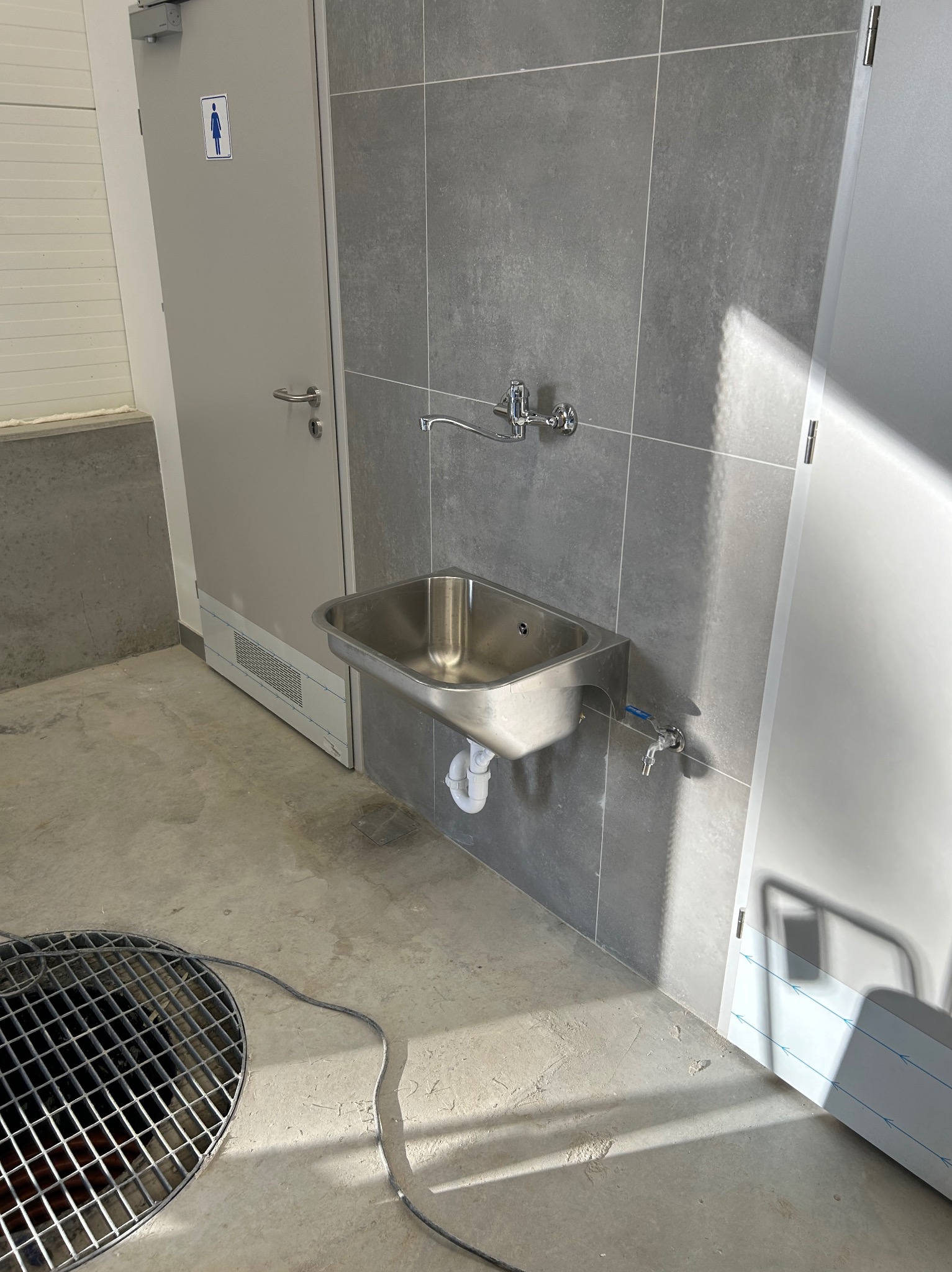 Stalowa umywalka zamontowana na szarej ścianie z płytek w publicznej toalecie. Widoczny odpływ podłogowy i rury. Drzwi z symbolem toalety damskiej.
