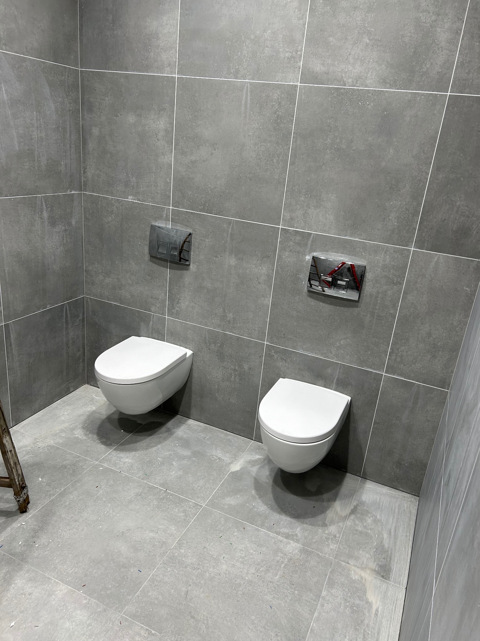 Dwa podwieszane, białe sedesy z chromowanymi przyciskami spłukującymi na szarej, kwadratowej ścianie z płytek. U dołu widoczny fragment drabiny.