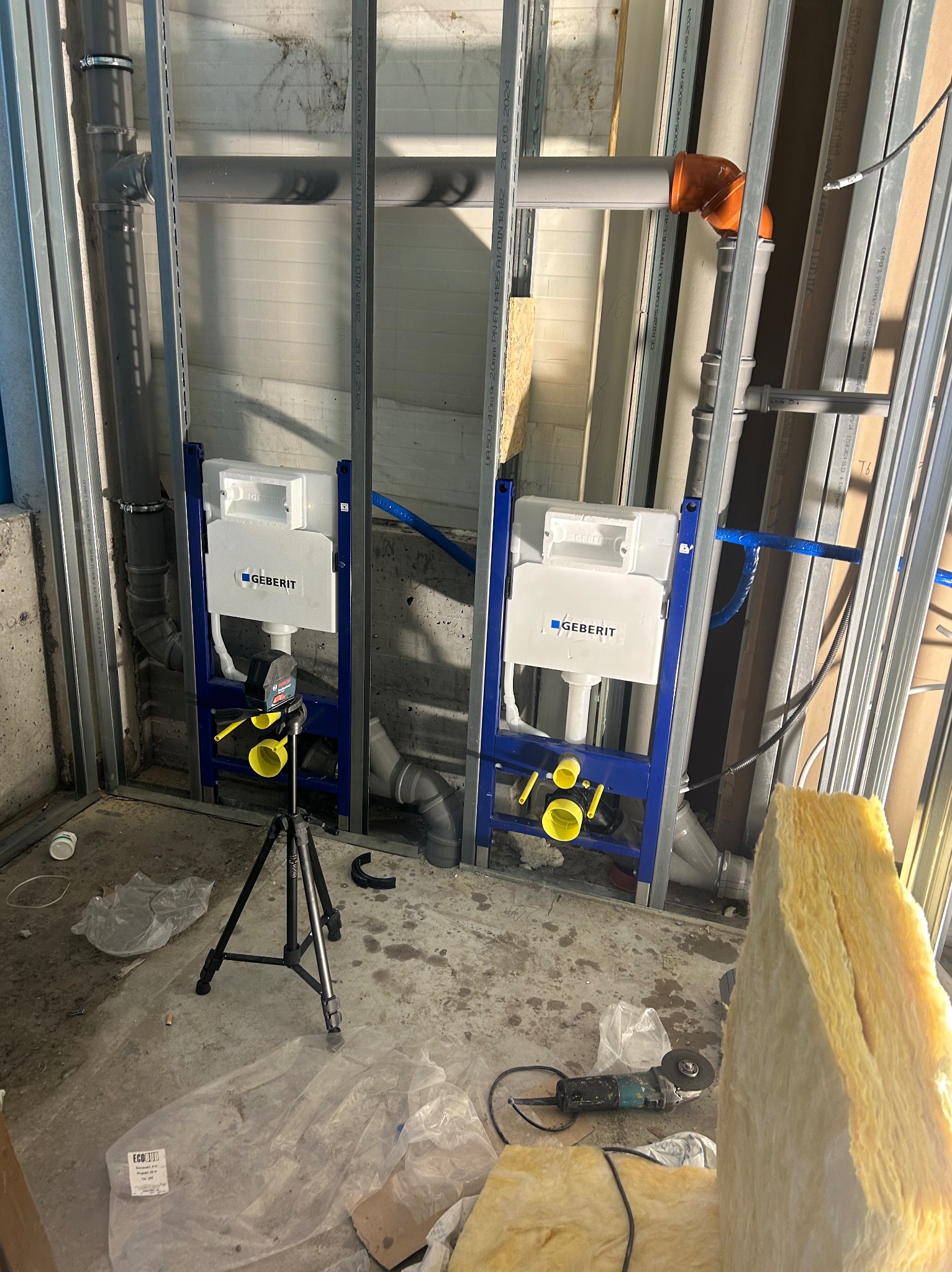 Stelaże podtynkowe Geberit zamontowane w metalowej konstrukcji, widoczne rury, izolacja i narzędzia. Prace instalacyjne w surowym wnętrzu.