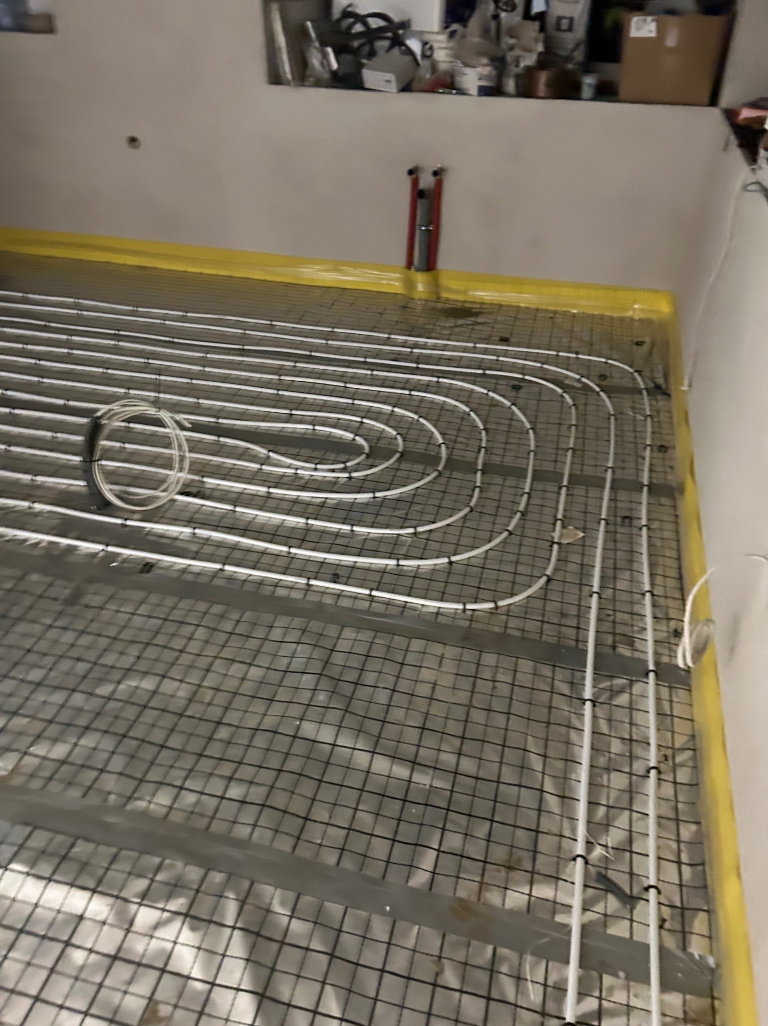 Instalacja ogrzewania podłogowego: białe rury ułożone w pętle na czarnej siatce, zabezpieczone żółtą folią przy ścianach. Widoczne elementy montażowe.