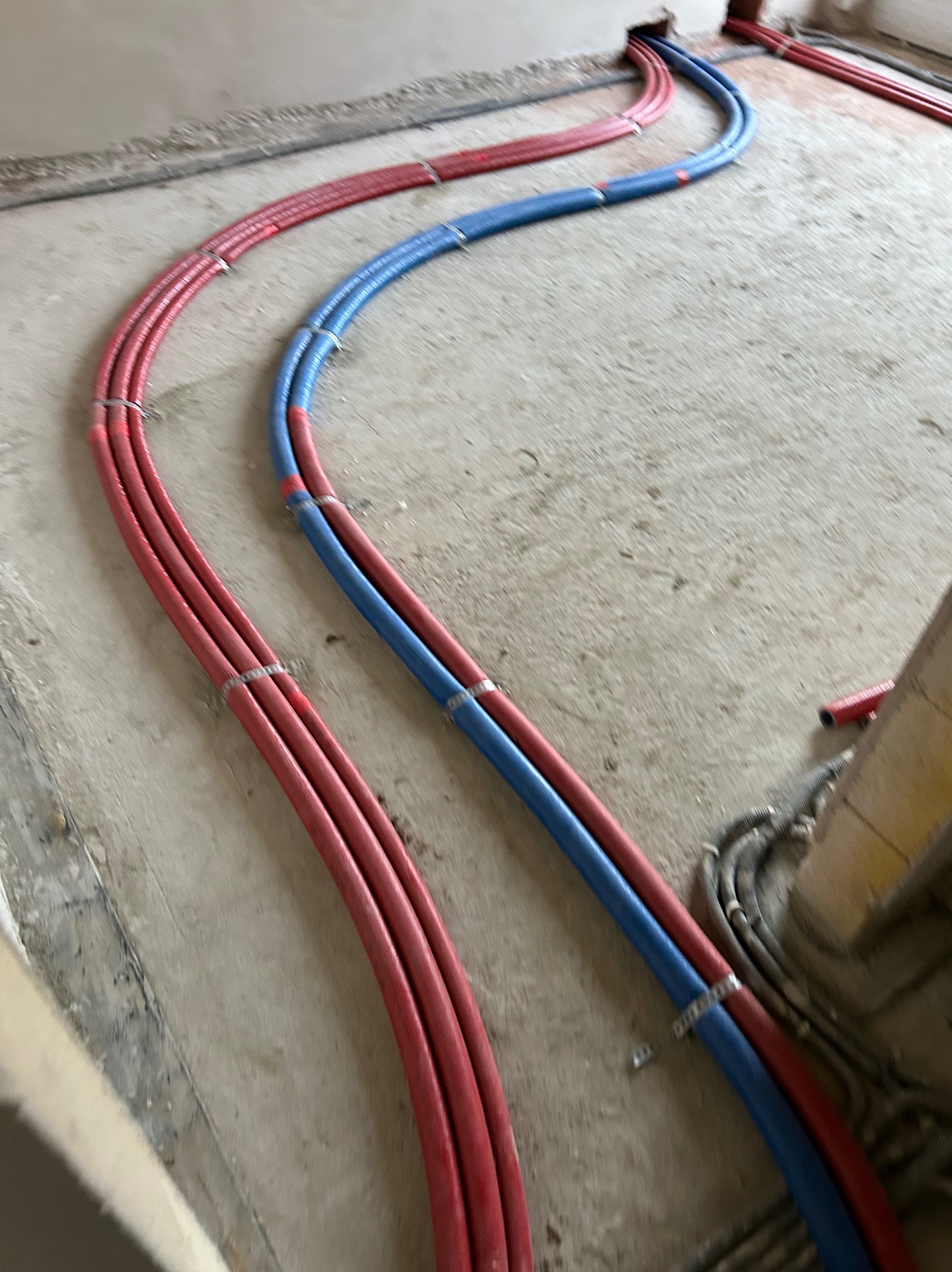 Instalacja ogrzewania podłogowego w trakcie montażu: czerwone i niebieskie rury ułożone na surowym betonie, zabezpieczone srebrnymi klamrami.