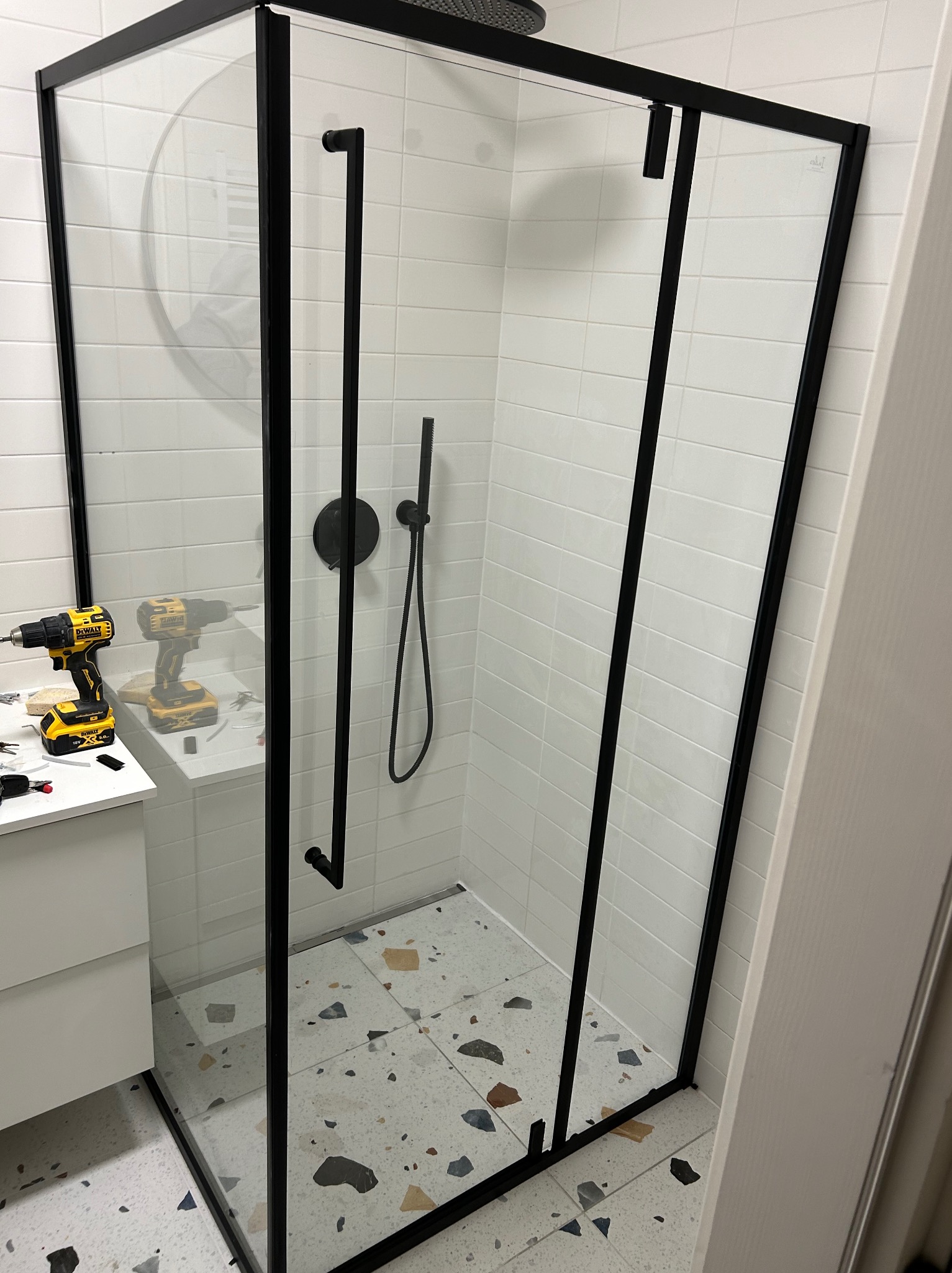 Nowoczesna kabina prysznicowa z czarnymi profilami, białe płytki i terazzo. W tle wiertarki sugerują montaż w trakcie realizacji.