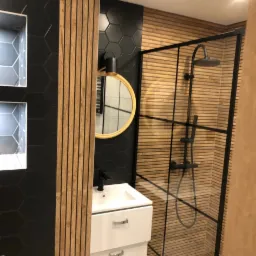 Nowoczesna łazienka z czarnymi heksagonalnymi płytkami, drewnianymi lamelami, białą szafką umywalkową z czarną baterią i kabiną prysznicową typu walk-in z czarnymi profilami.