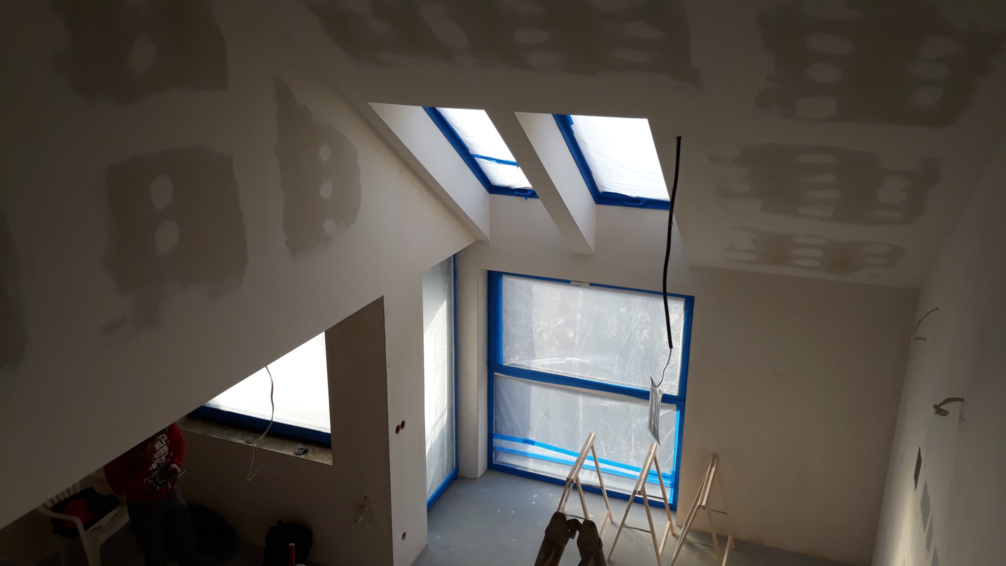 Poddasze w trakcie prac przygotowawczych: okna zabezpieczone folią i taśmą, ściany i sufit z widocznymi łatami po szpachlowaniu, drabina malarska w pomieszczeniu.