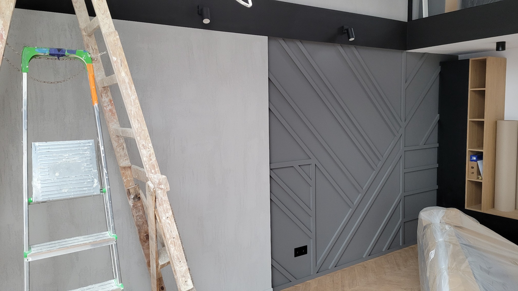 Szara ściana z dekoracyjnymi listwami i gładzią, drabina malarska z resztkami farby, podłoga parkietowa, meble zabezpieczone folią, sufit z oświetleniem punktowym.