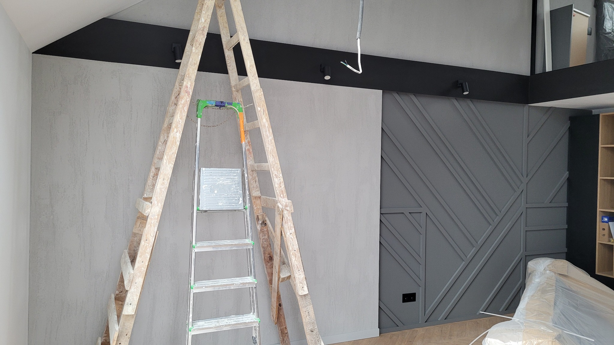 Dwie drabiny opierają się o ścianę z szarą dekoracyjną strukturą i ozdobną szarą ścianą z geometrycznym wzorem, widoczne oświetlenie punktowe i fragment podłogi.