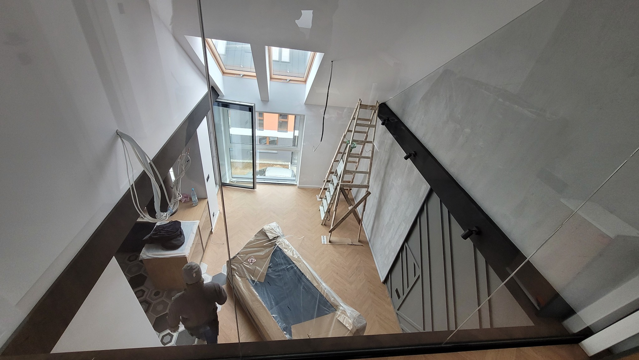 Wysoki kąt widzenia na wnętrze w trakcie wykańczania: drewniana drabina oparta o gładką, szarą ścianę z dekoracyjnymi pionowymi listwami, okna dachowe w suficie, podłoga z parkietu w jodełkę...