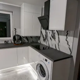 Nowoczesna kuchnia z białymi, lakierowanymi szafkami, czarnym blatem i marmurowym backsplash'em, zintegrowana z pralką, oświetlenie LED pod szafkami, szare płytki podłogowe.