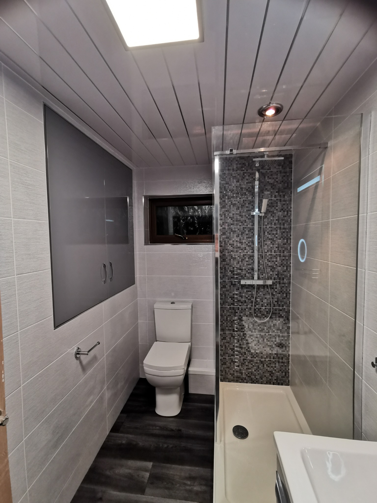 Nowoczesna łazienka z szarymi płytkami imitującymi drewno na ścianach, ciemną podłogą, kabiną prysznicową z mozaiką i szafką w zabudowie.