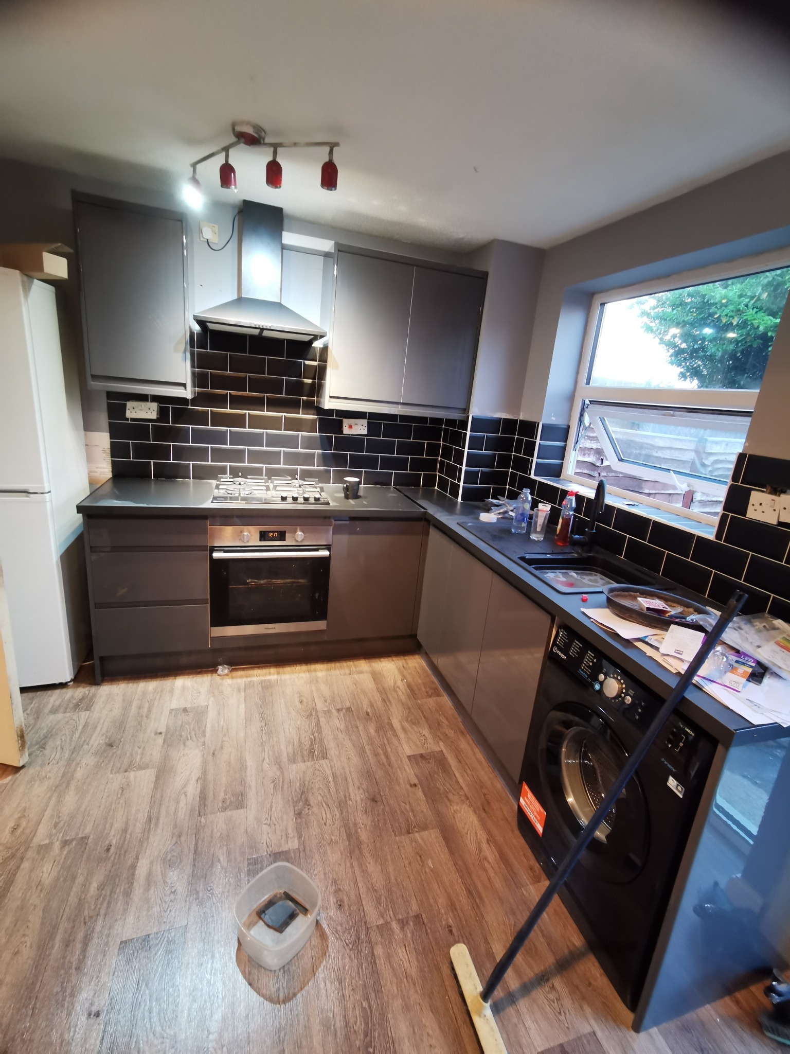 Szara kuchnia z czarnymi płytkami, widoczna pralka pod blatem, okno z widokiem na zieleń, podłoga panelowa i mop oparty o pralkę.