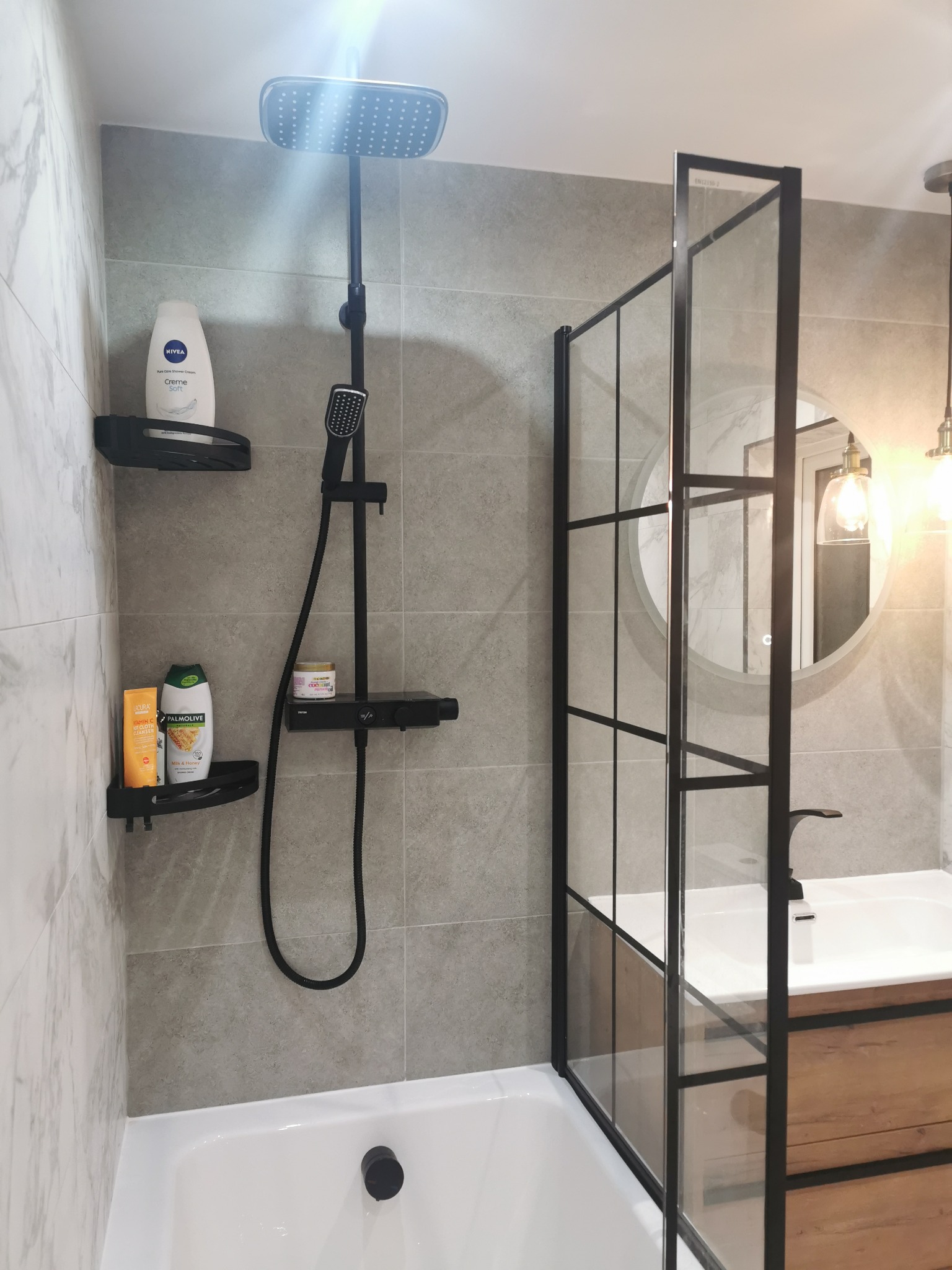 Nowoczesna łazienka z kabiną prysznicową typu walk-in z czarnymi profilami, szarymi płytkami imitującymi marmur, czarną armaturą i drewnianą szafką pod umywalką, oświetlona ciepłym światłem.