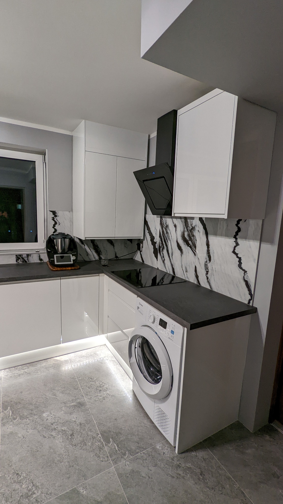 Nowoczesna kuchnia z białymi, lakierowanymi szafkami, czarnym blatem i marmurowym backsplash'em, zintegrowana z pralką, oświetlenie LED pod szafkami, szare płytki podłogowe.
