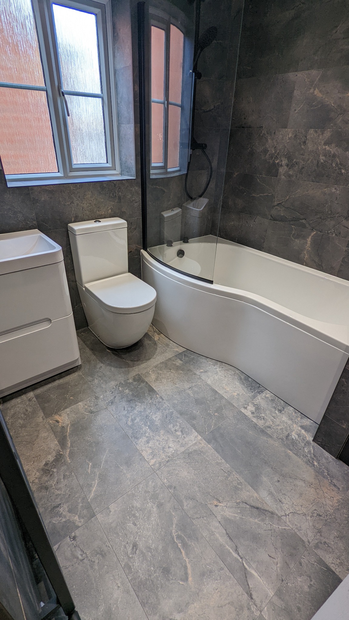 Nowoczesna łazienka wykończona szarymi płytkami imitującymi marmur, z białą wanną z parawanem, toaletą i umywalką z szafką. Widoczne okno z szybą ornamentową. Czarna armatura prysznicowa.