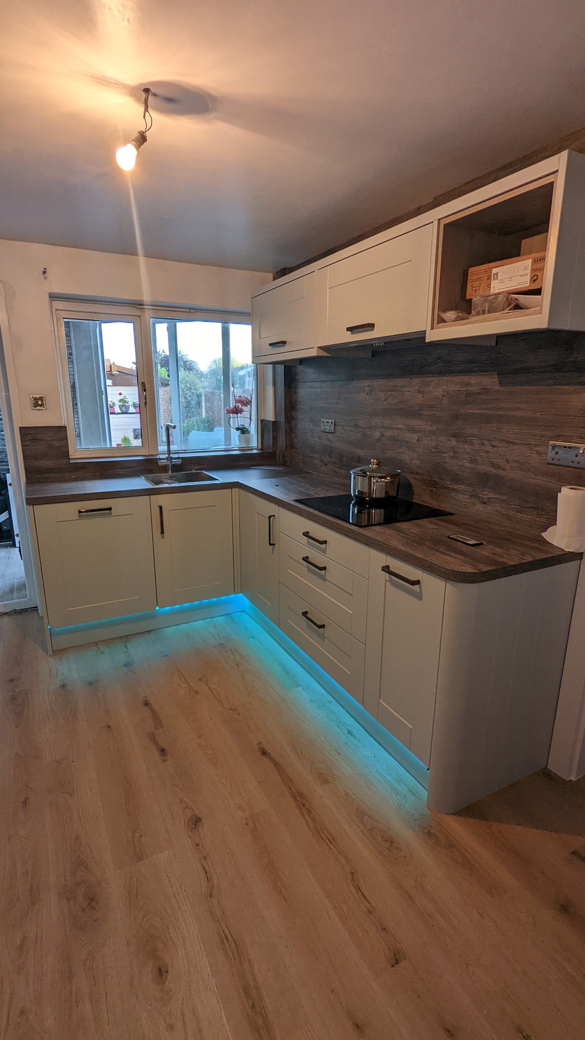 Jasna kuchnia z drewnianym blatem i podświetleniem LED pod szafkami, na blacie indukcyjnym stoi garnek.