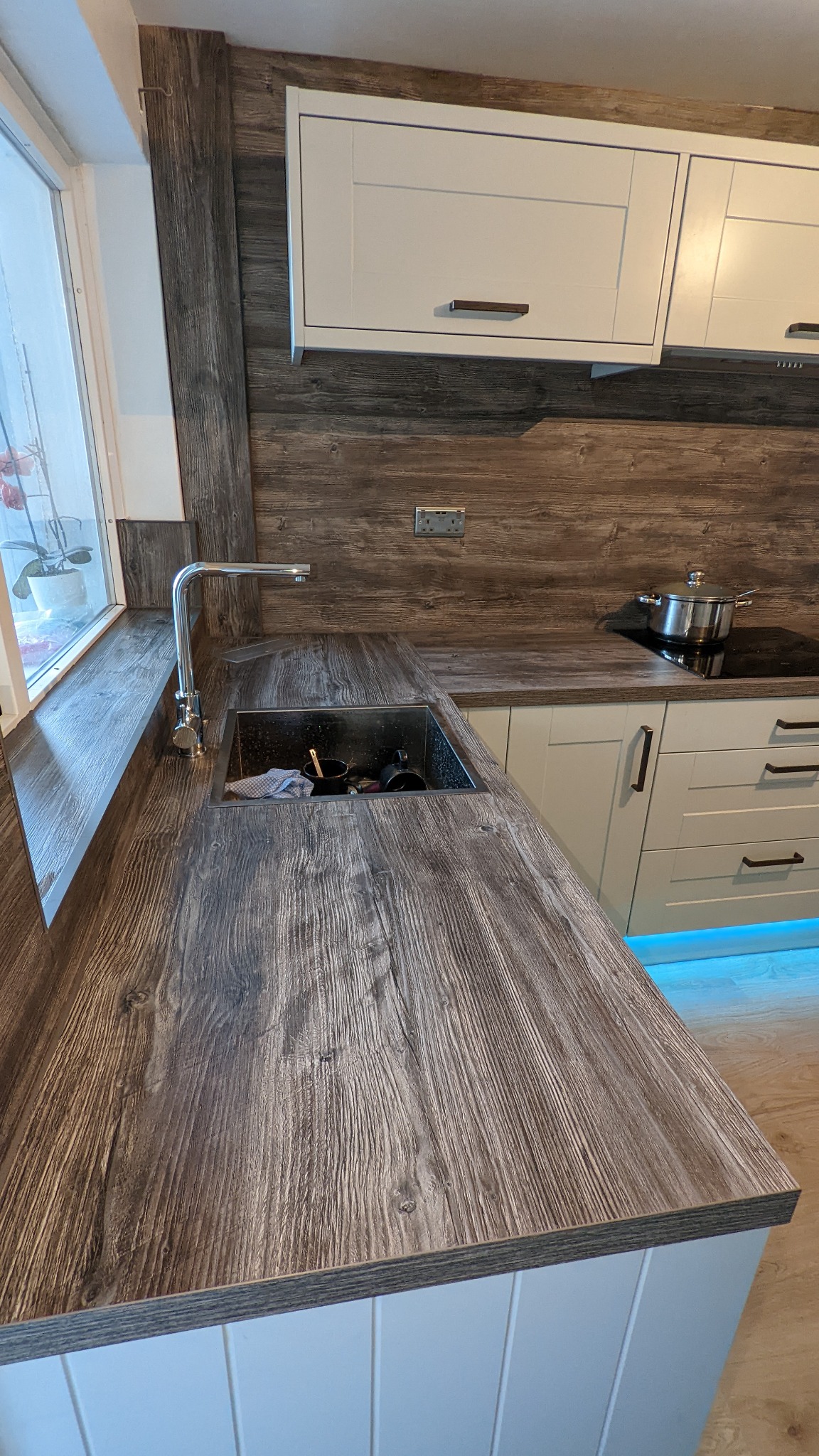 Nowoczesna kuchnia z drewnianym blatem, zlewozmywakiem ze stali nierdzewnej i białymi szafkami, podświetleniem LED pod szafkami dolnymi oraz panelem ściennym imitującym drewno.