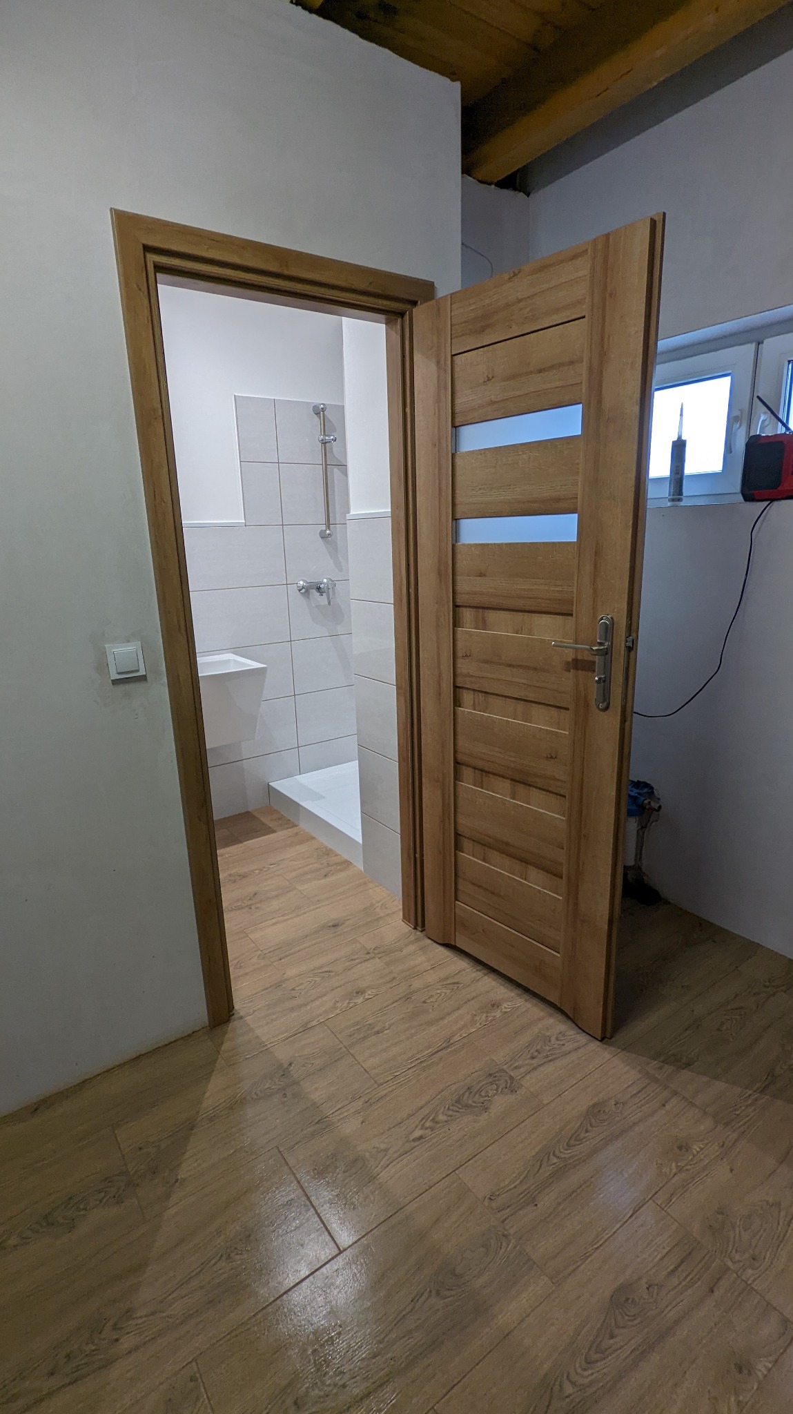 Otwarta drewniana drzwi z przeszkleniami, widok na wyremontowaną łazienkę z białymi płytkami, kabiną prysznicową i wiszącą umywalką, podłoga wyłożona panelami imitującymi drewno.