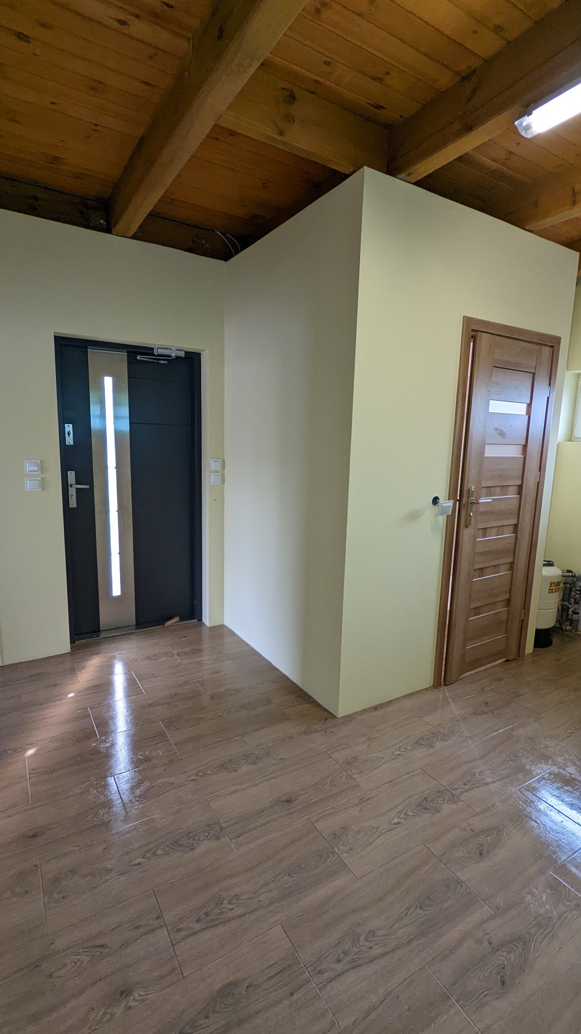 Jasne wnętrze z drewnianym sufitem, brązowymi płytkami imitującymi drewno na podłodze, dwojgiem drzwi: ciemnymi z pionowym przeszkleniem i jasnymi drewnianymi z poziomymi wstawkami, świeżo...